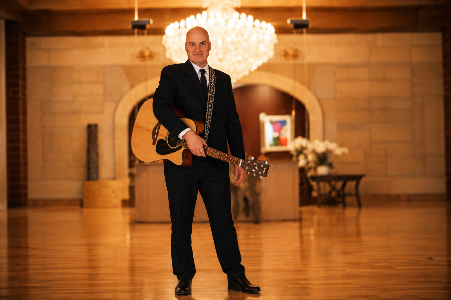 A man in a black suit holding an acoustic guitar inside a warmly lit room with wooden floors and a chandelier, standing in front of an arched wall with a framed painting and bouquet of flowers on a table.
