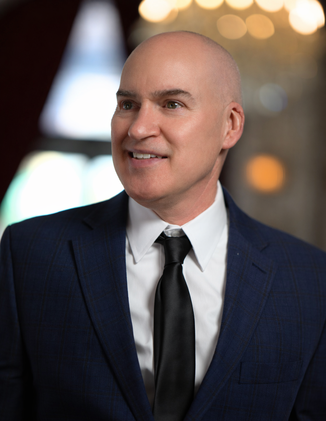 A man with a shaved head dressed in a formal dark checkered suit, white shirt, and black tie smiling in an indoor setting with blurred warm lights in the background.