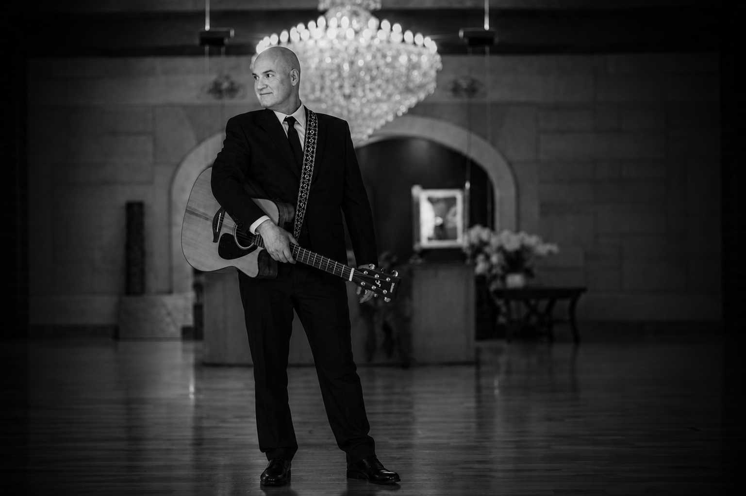 A bald man dressed in a tuxedo, holding an acoustic guitar, standing in a luxurious room with a chandelier and floral arrangements in the background.