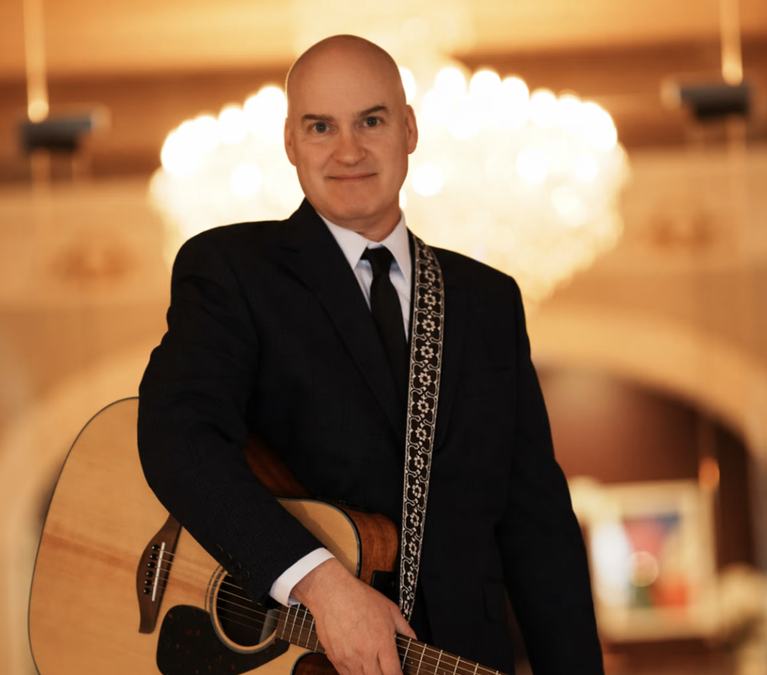 A man in a suit holding an acoustic guitar, standing in a warmly lit, elegant interior with a chandelier in the background.