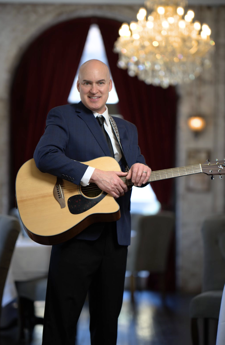 A man in a navy blue suit playing an acoustic guitar in a room with a chandelier, red curtains, and stone walls.