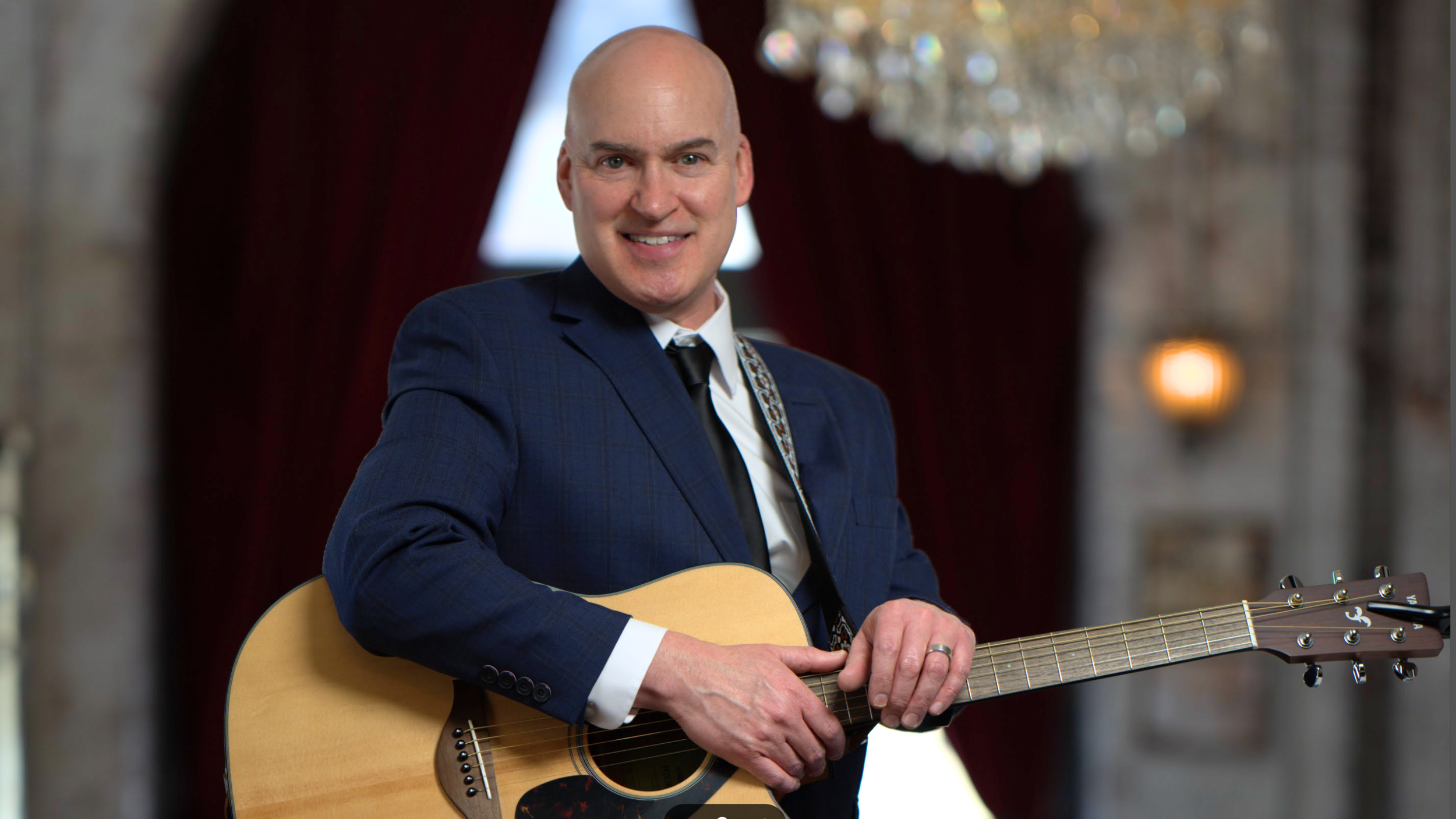A man in a blue suit playing an acoustic guitar in a room with a chandelier and draped curtains.