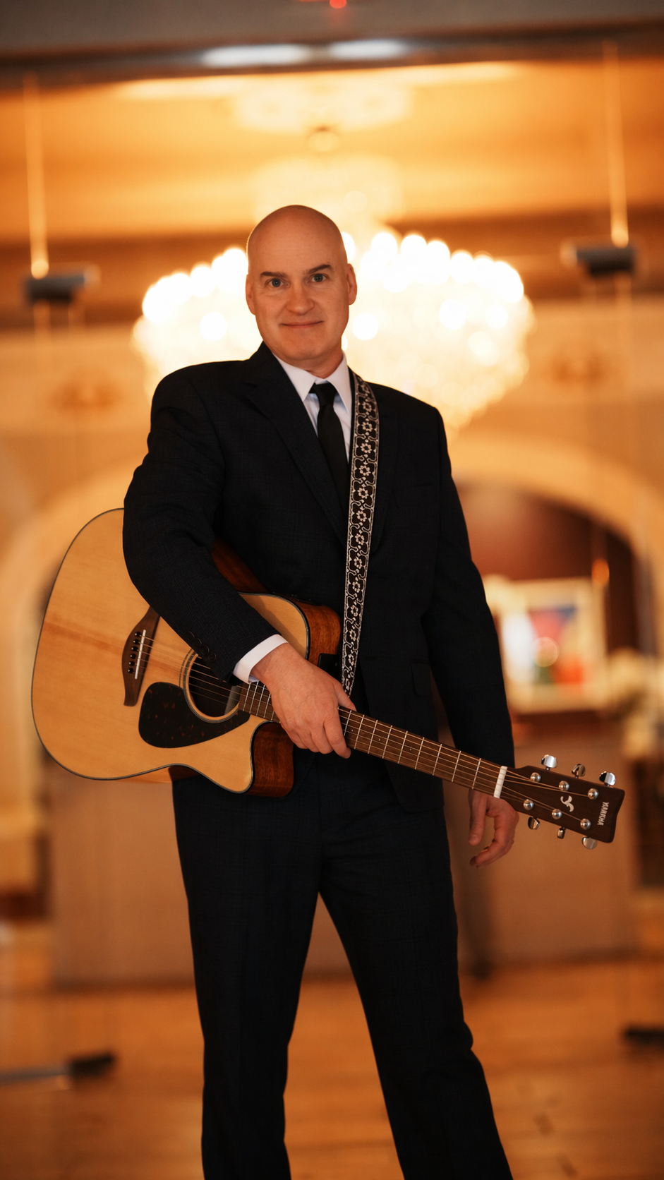 A man in a black suit holding an acoustic guitar in a warmly lit room with a chandelier in the background.