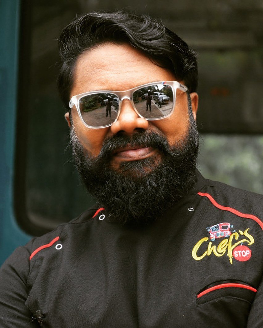 A man with a dark beard and mustache wearing reflective sunglasses and a black shirt with red and yellow embroidery, standing outdoors with a blurred background.