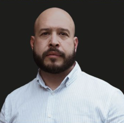 A man with a bald head, beard, and mustache wearing a light blue button-up shirt, posing against a black background.