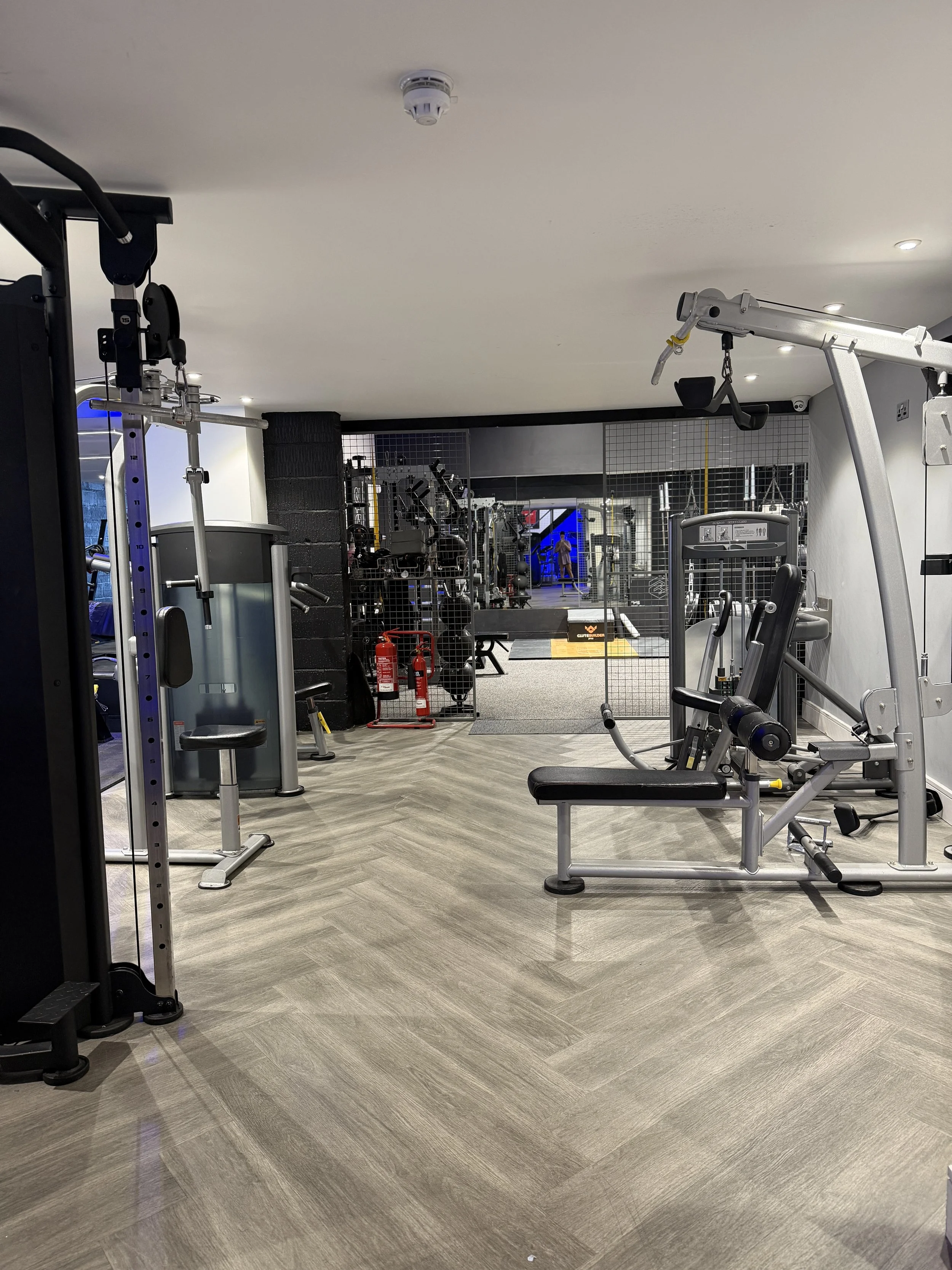 Interior of a gym with various exercise equipment, including weight machines and free weights, with a mirror at the back reflecting the gym space.