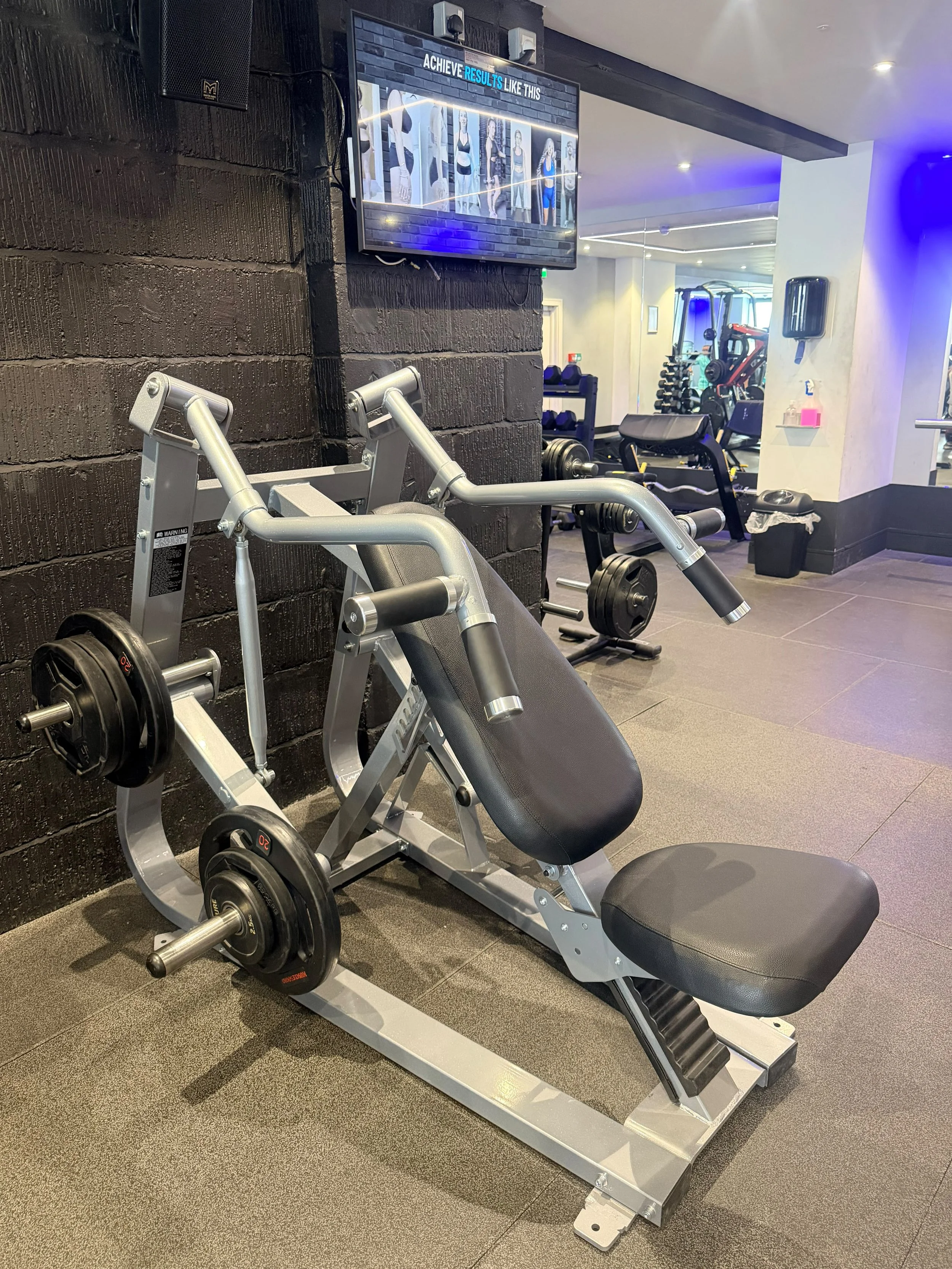 Workout gym with a leg press machine in the foreground, dumbbell rack in the background, and television mounted on a black brick wall displaying fitness results.