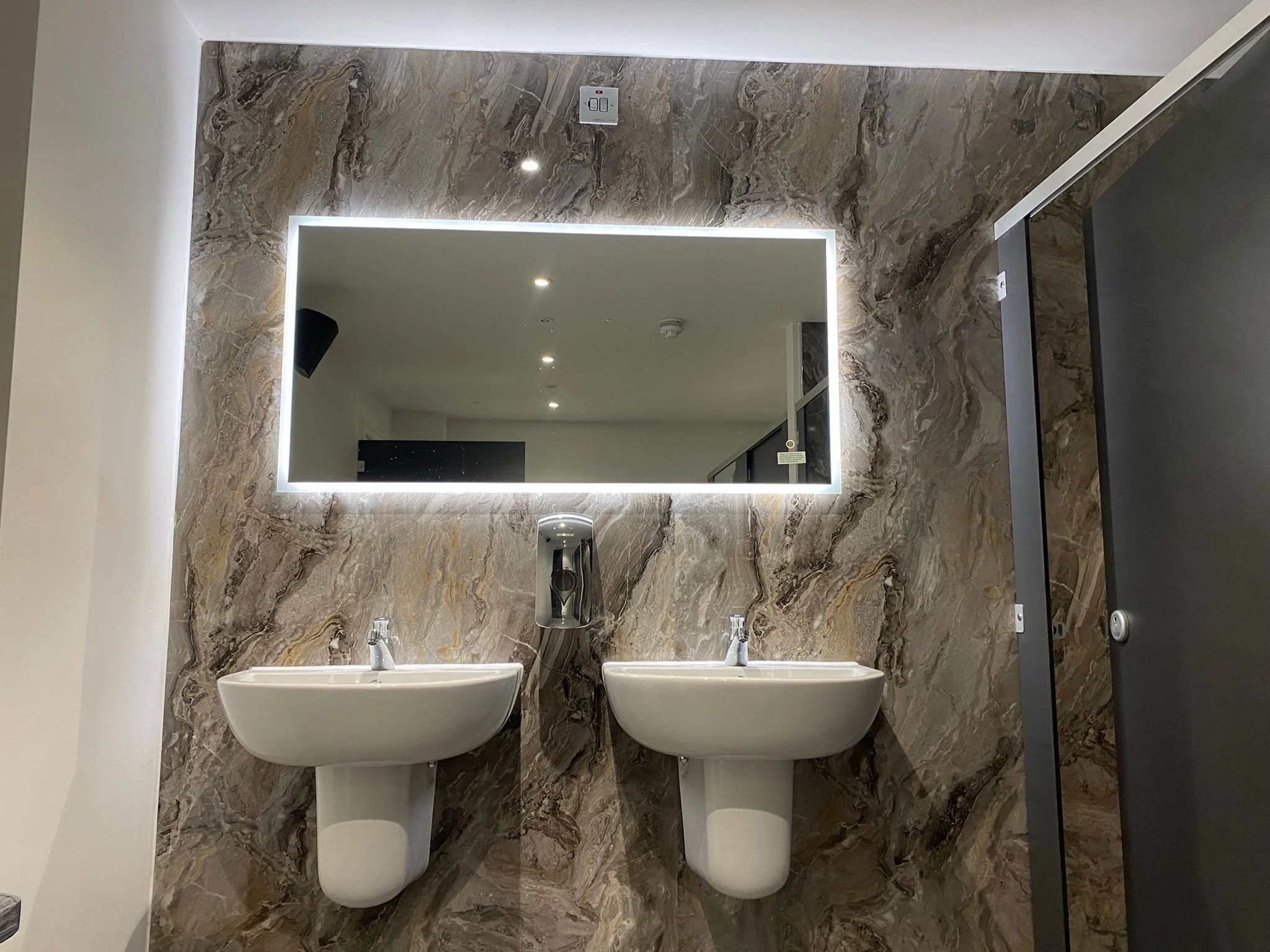 Bathroom with marble walls, two white wall-mounted sinks, and a large rectangular mirror illuminated by LED lighting.