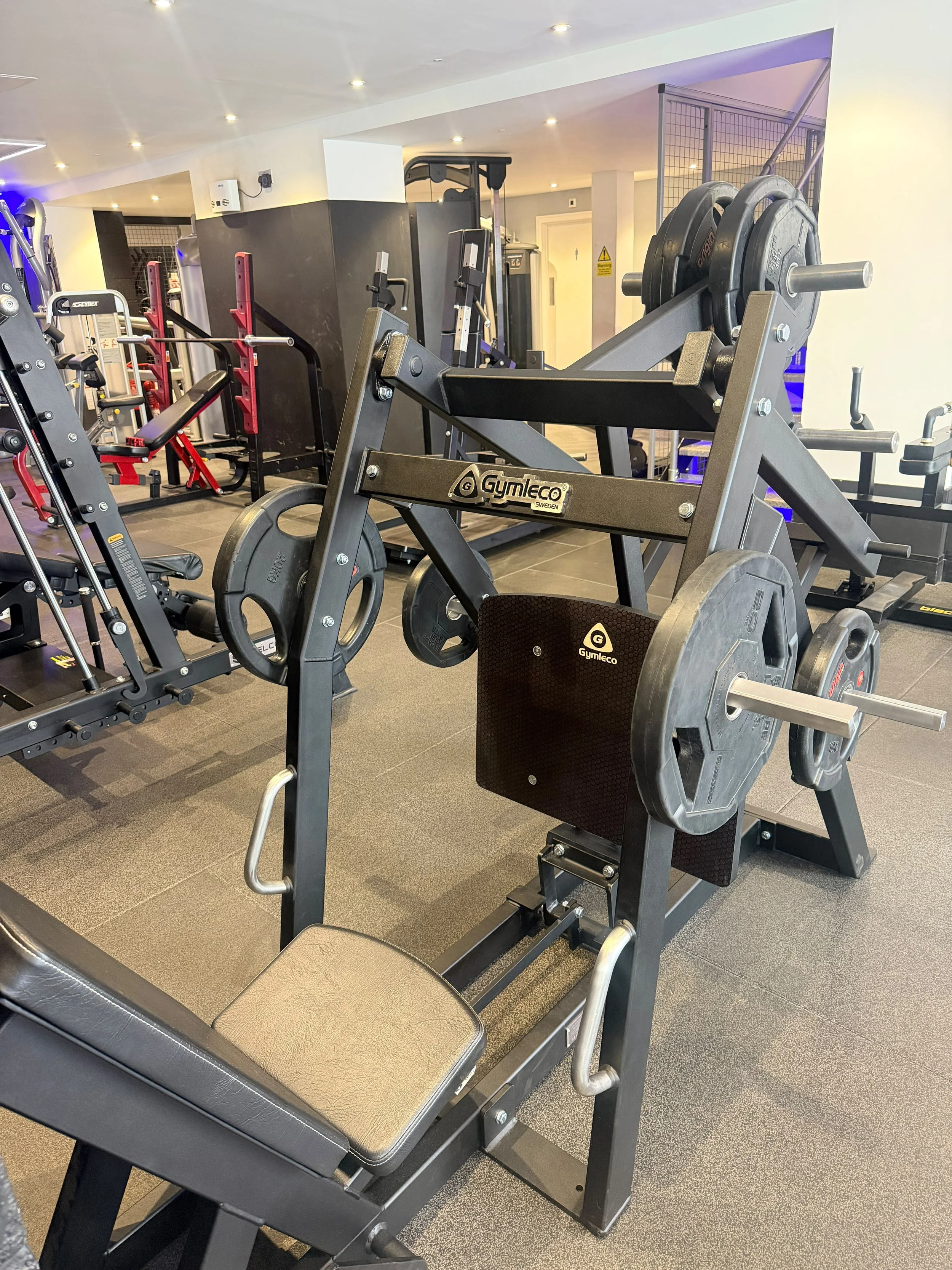 Gym equipment, including weightlifting machines and benches, in a well-lit fitness center.