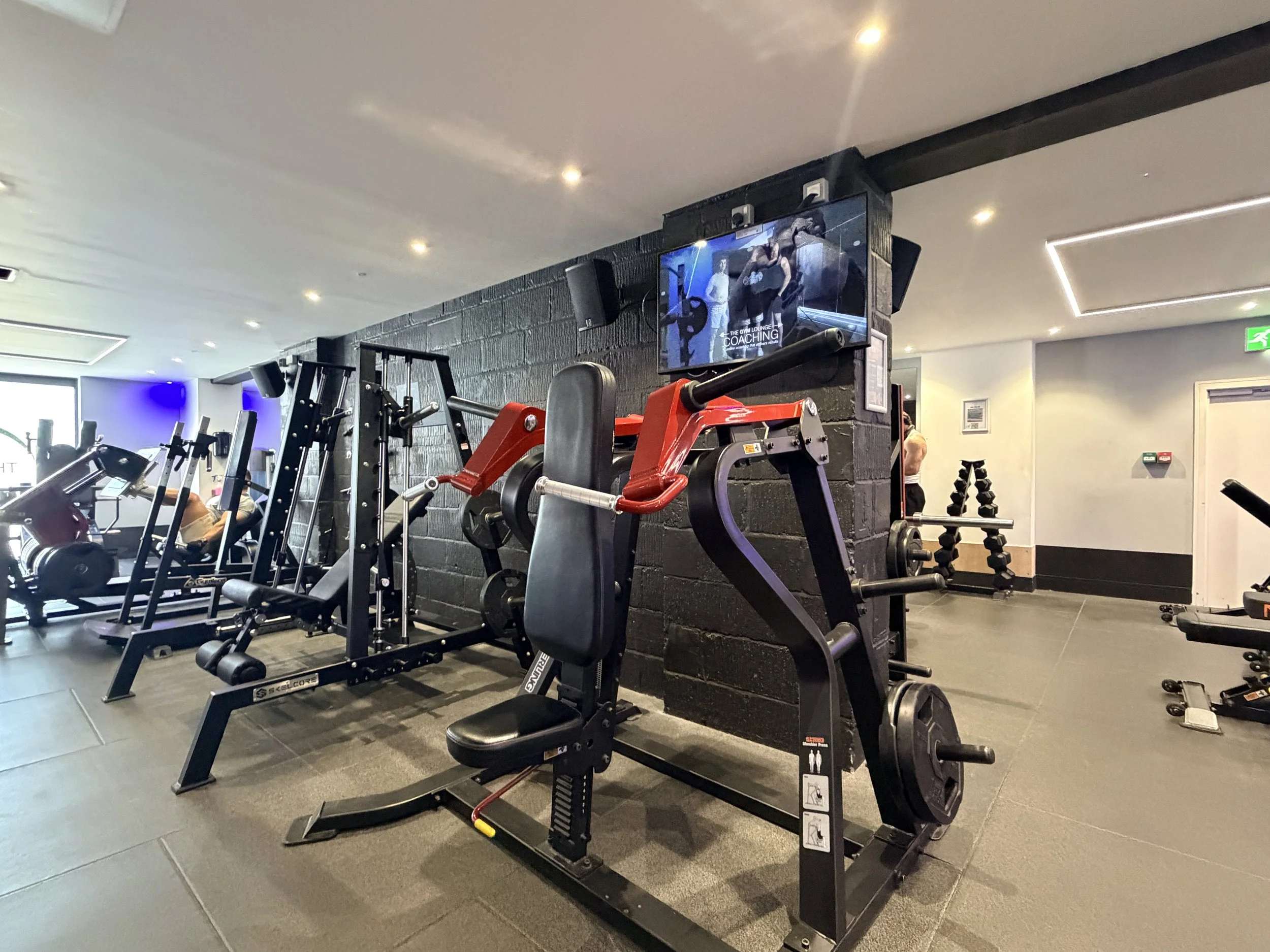 Interior of a gym with various exercise equipment, including weight machines and a weight bench, and a television mounted on a brick wall showing people working out.