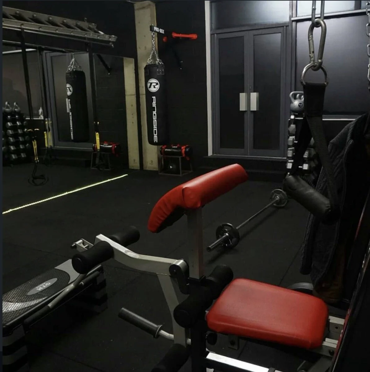 Home gym with black flooring, including a red and black weight bench, punching bags, and various exercise equipment.