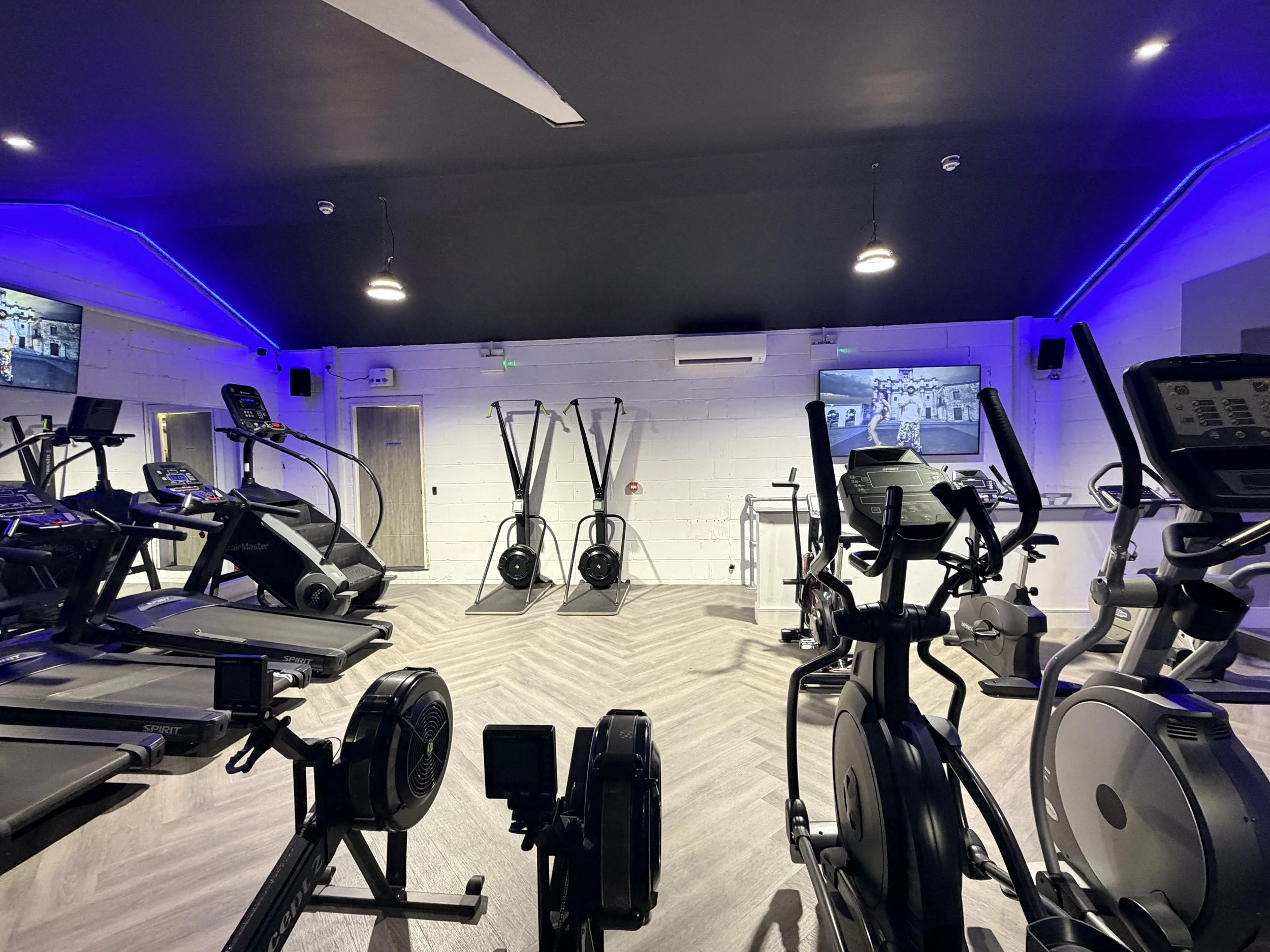 A gym with various cardio machines including treadmills, stationary bikes, and ellipticals. The room has white brick walls, wooden flooring, and blue accent lighting along the ceiling. Two large monitors display a fitness video, and fitness equipment