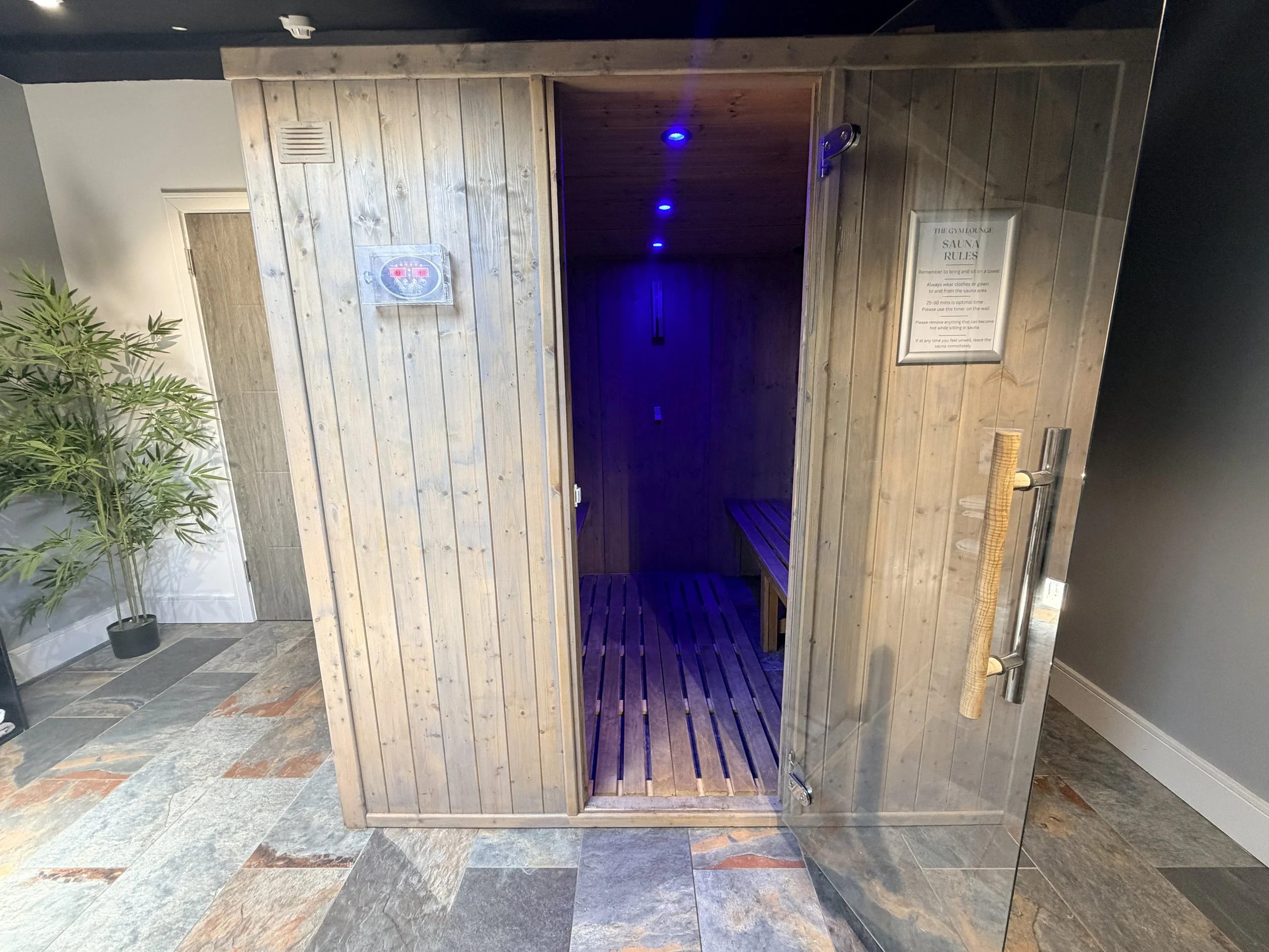 Wooden sauna room with blue lighting, a vent on the exterior wall, a sign with sauna rules, and a plant nearby, located indoors with slate tile flooring.