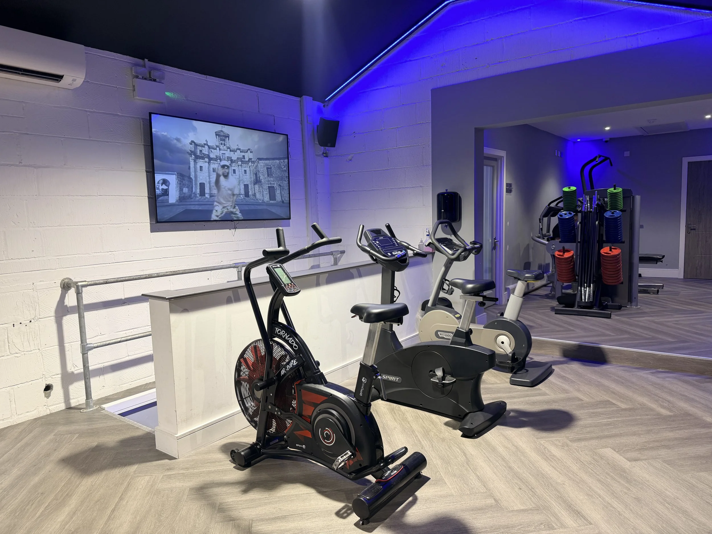 Gym with exercise bikes, weight rack, a TV showing a building, and blue LED lighting.