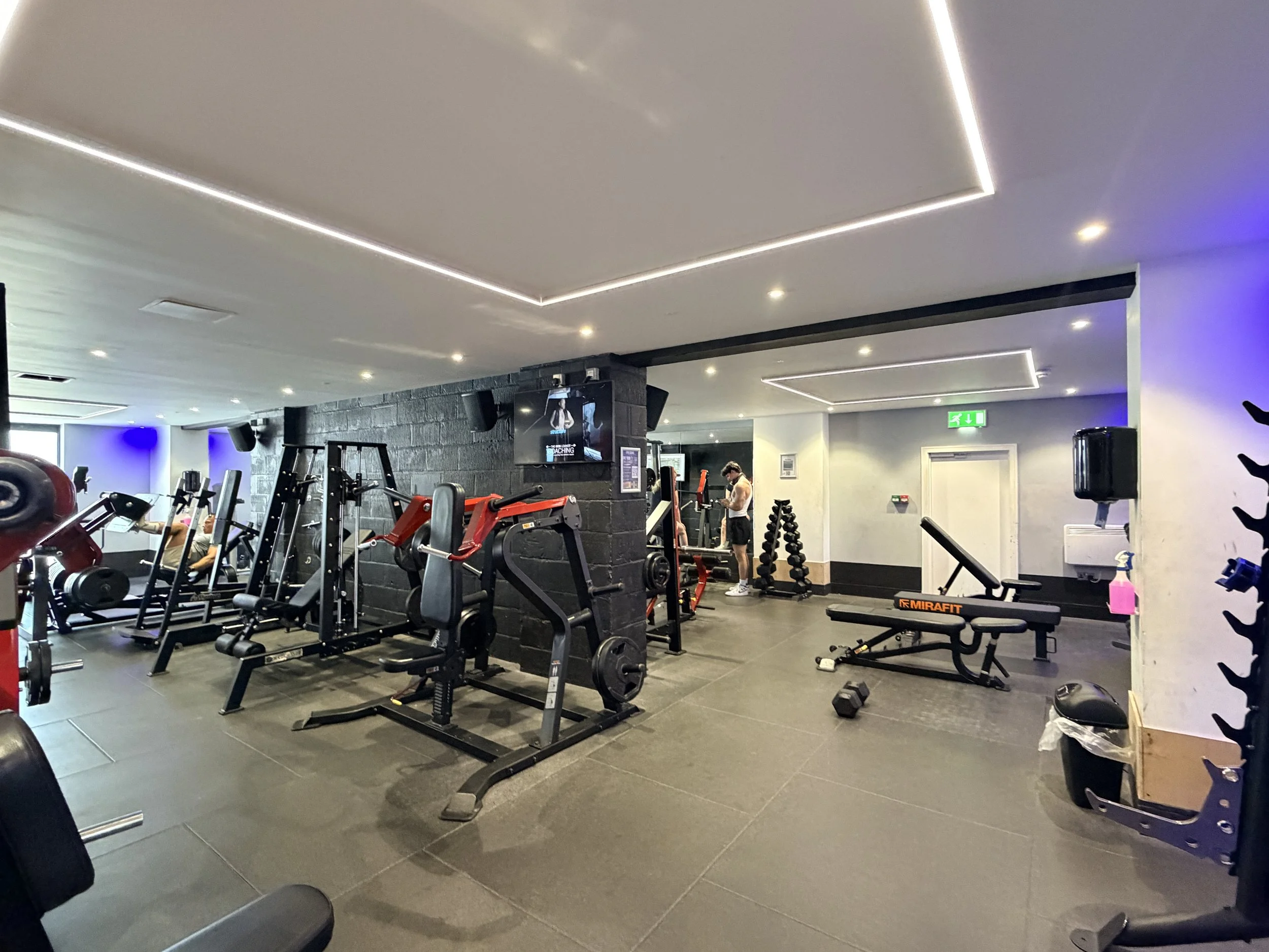 Interior of a gym with various exercise equipment, including weight machines, free weights, and benches, with people working out and a digital screen on a black brick wall.