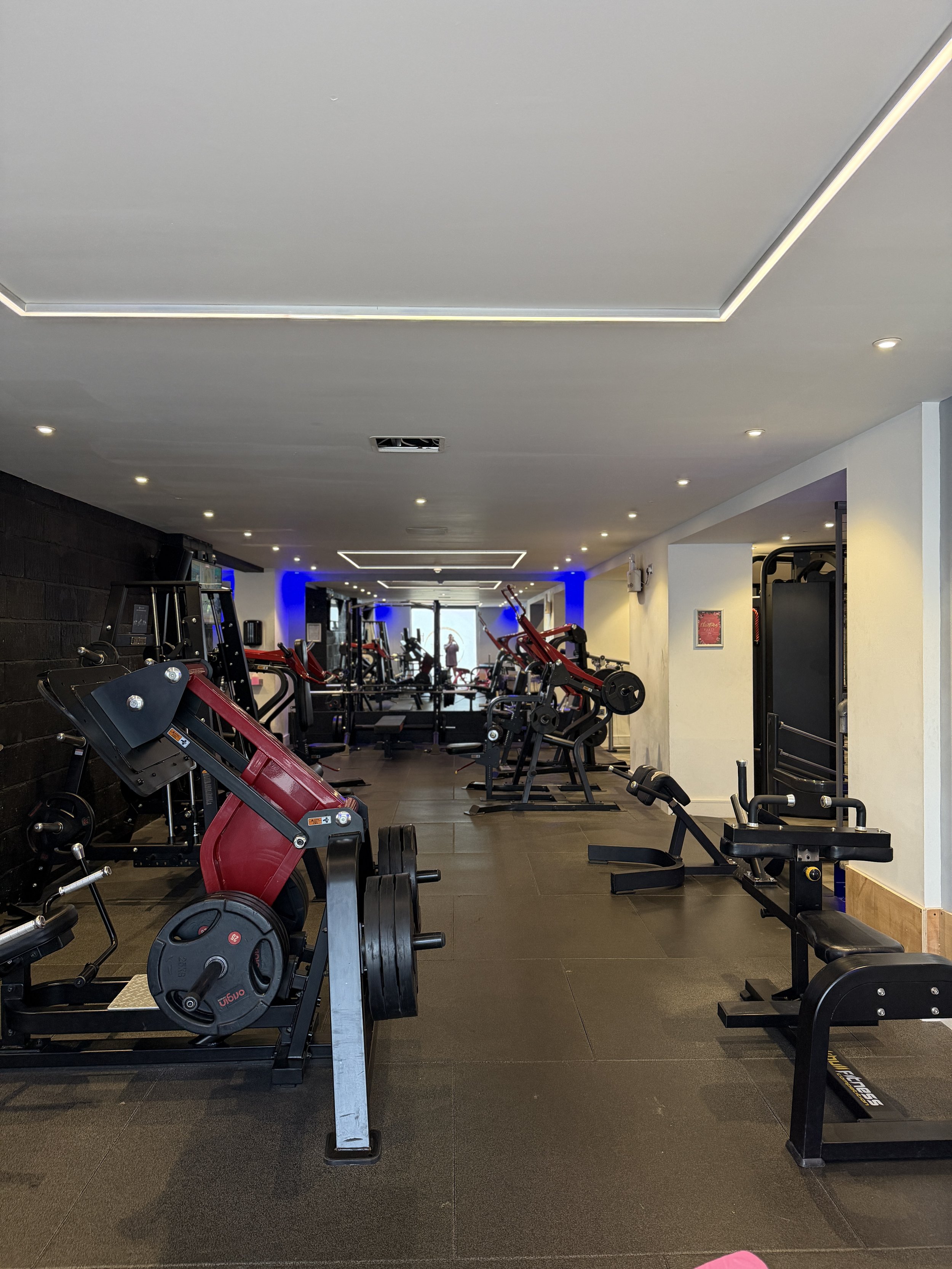 A fitness gym with various workout equipment including weights, benches, and cardio machines, illuminated by ceiling lights and a strip of LED lighting.