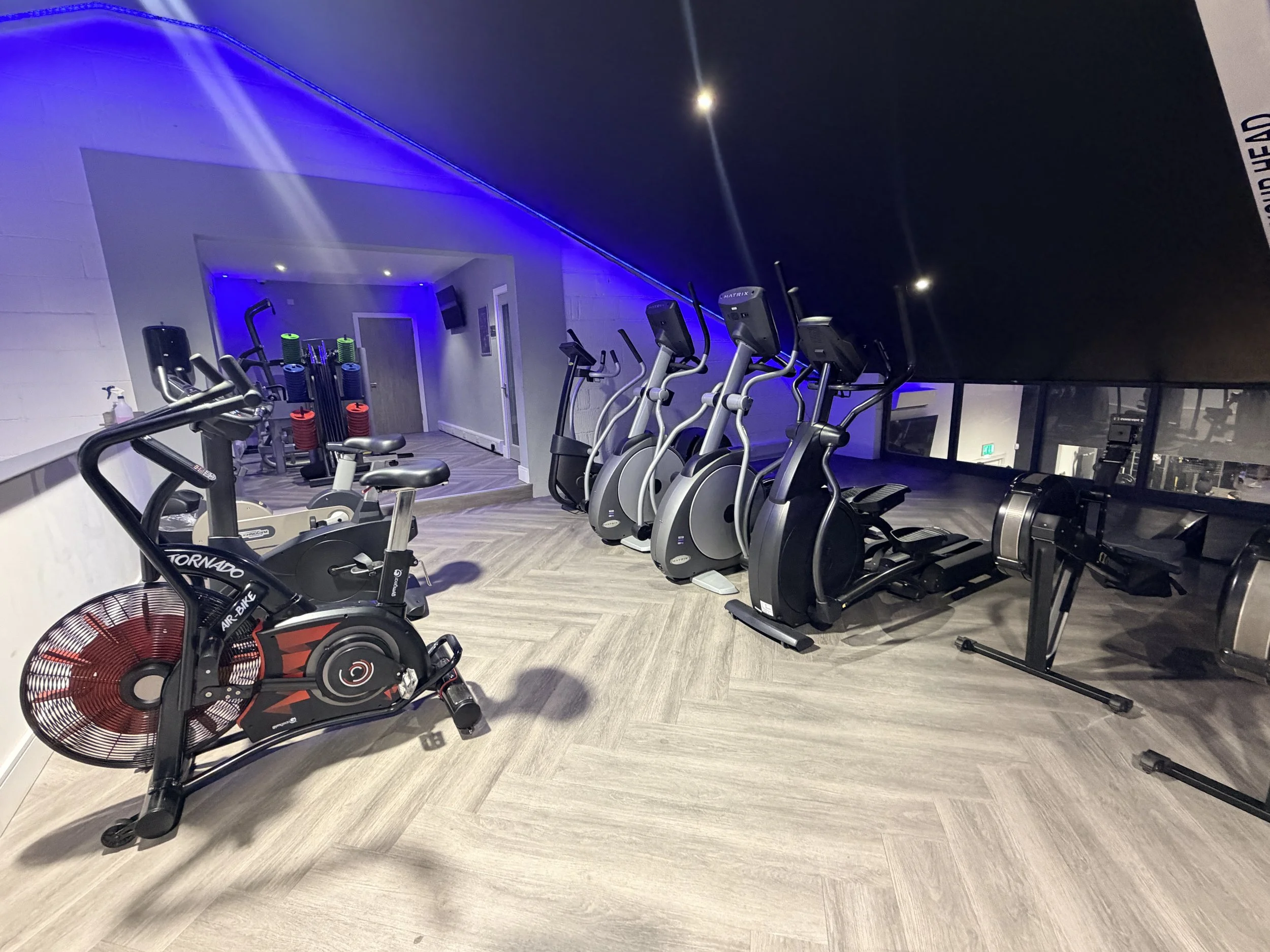 Interior of a gym with exercise equipment such as stationary bikes and elliptical machines, illuminated with blue lighting.