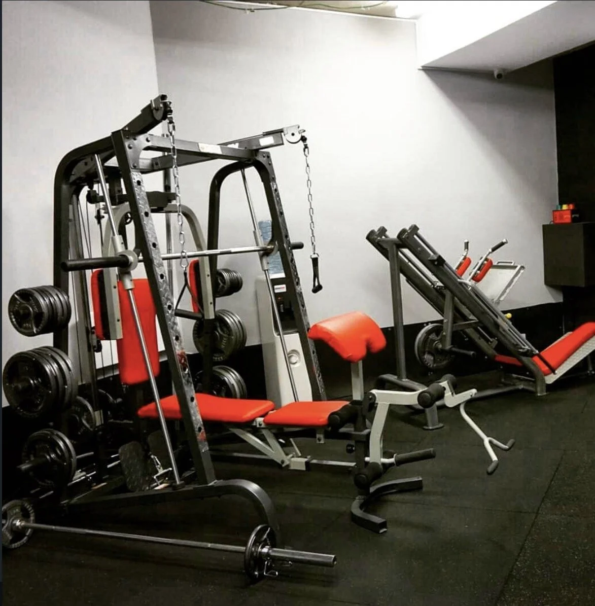 Gym equipment in a fitness center, including weight machines, dumbbells, a weight plate, a bench with red padding, and a water bottle on the machine in the background.
