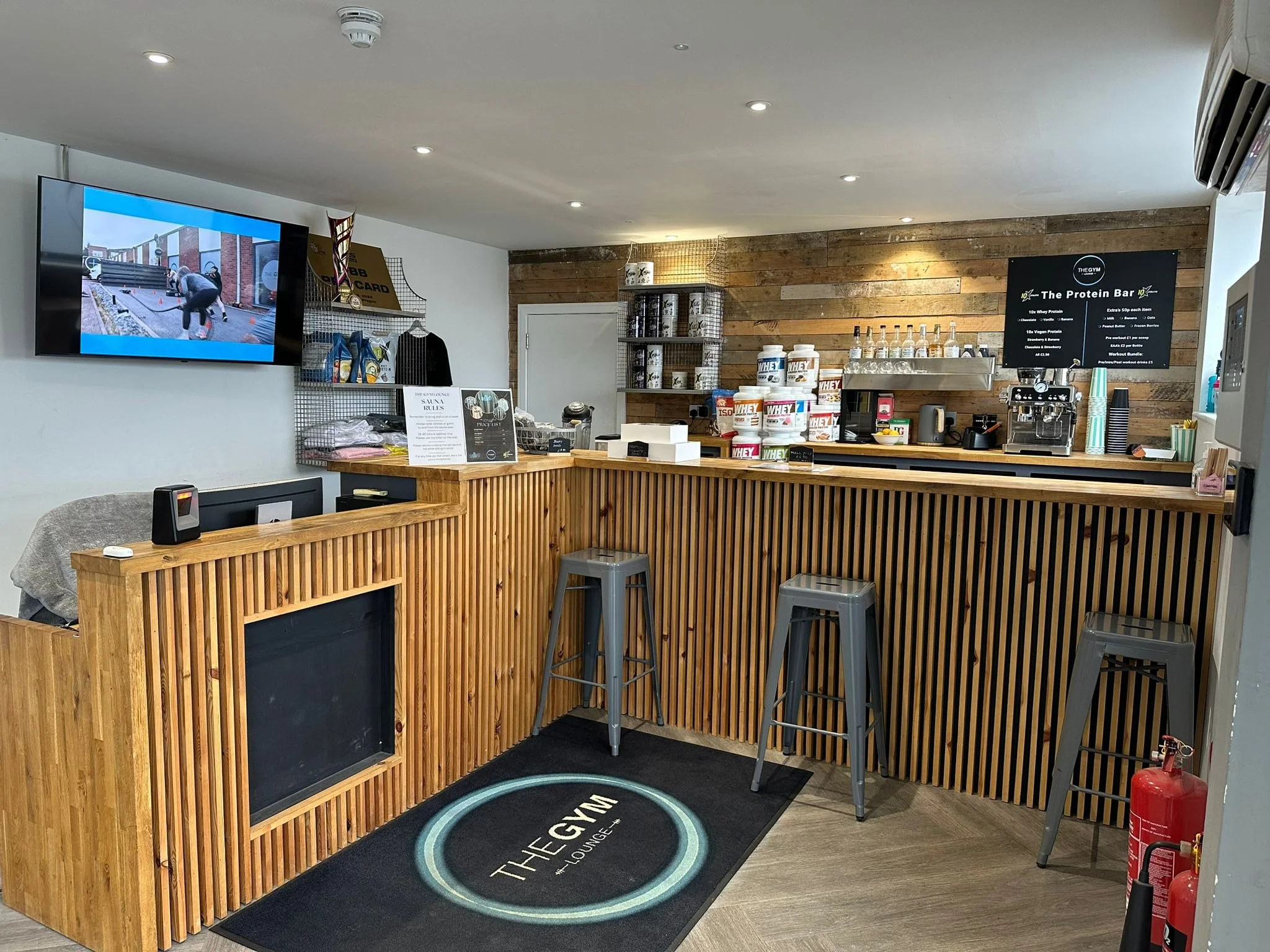 Interior of a fitness lounge area with wooden counter, a TV on the wall displaying outdoor exercise, shelves with protein supplements, a sign about gym rules, and a black mat with a gym brand logo.