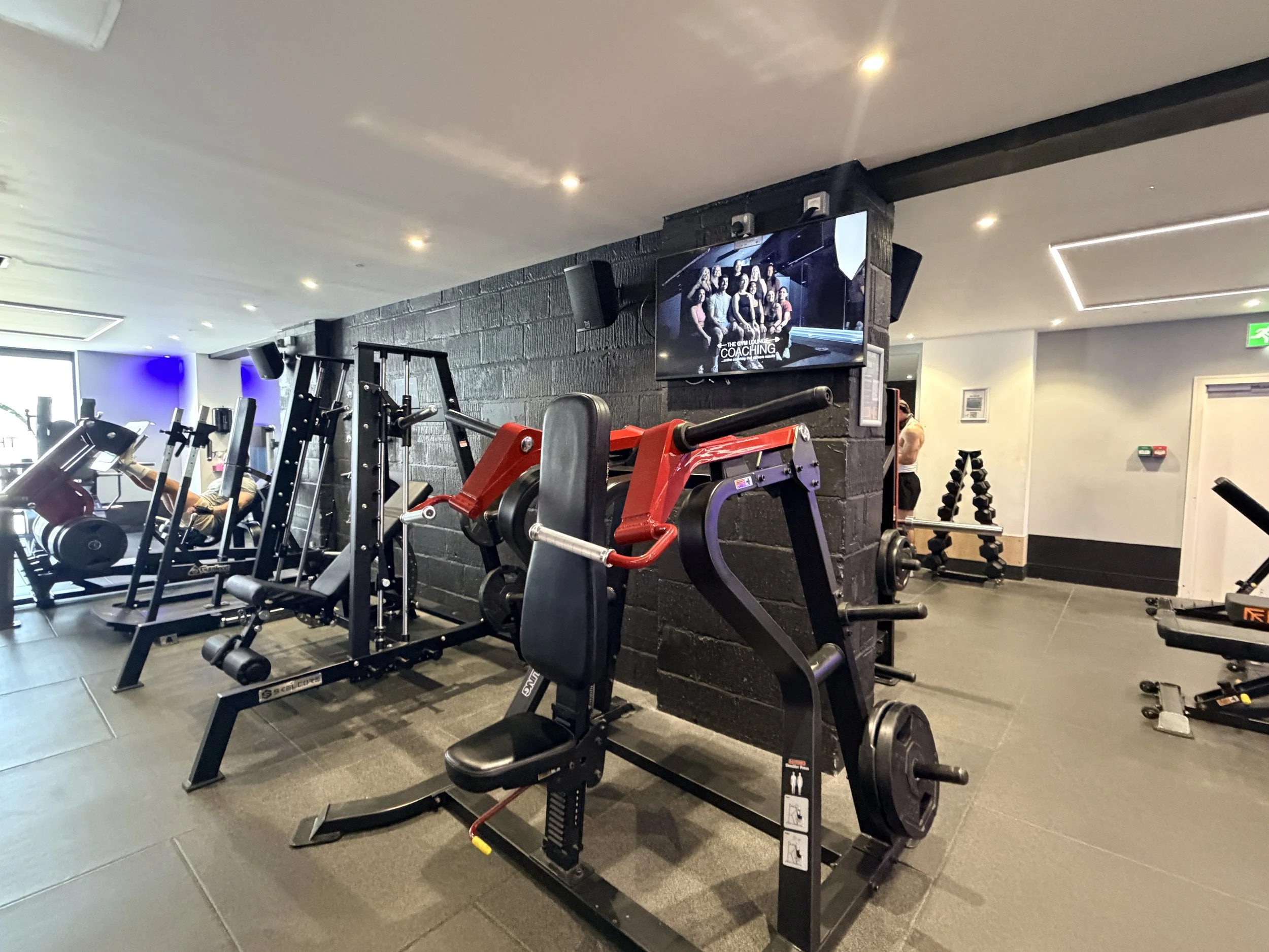 Interior of a gym with various workout equipment including weight machines and free weights, a wall-mounted screen displaying a group photo of people and the text 'The Gym Lounge Coaching', black stone walls, and ceiling lights.