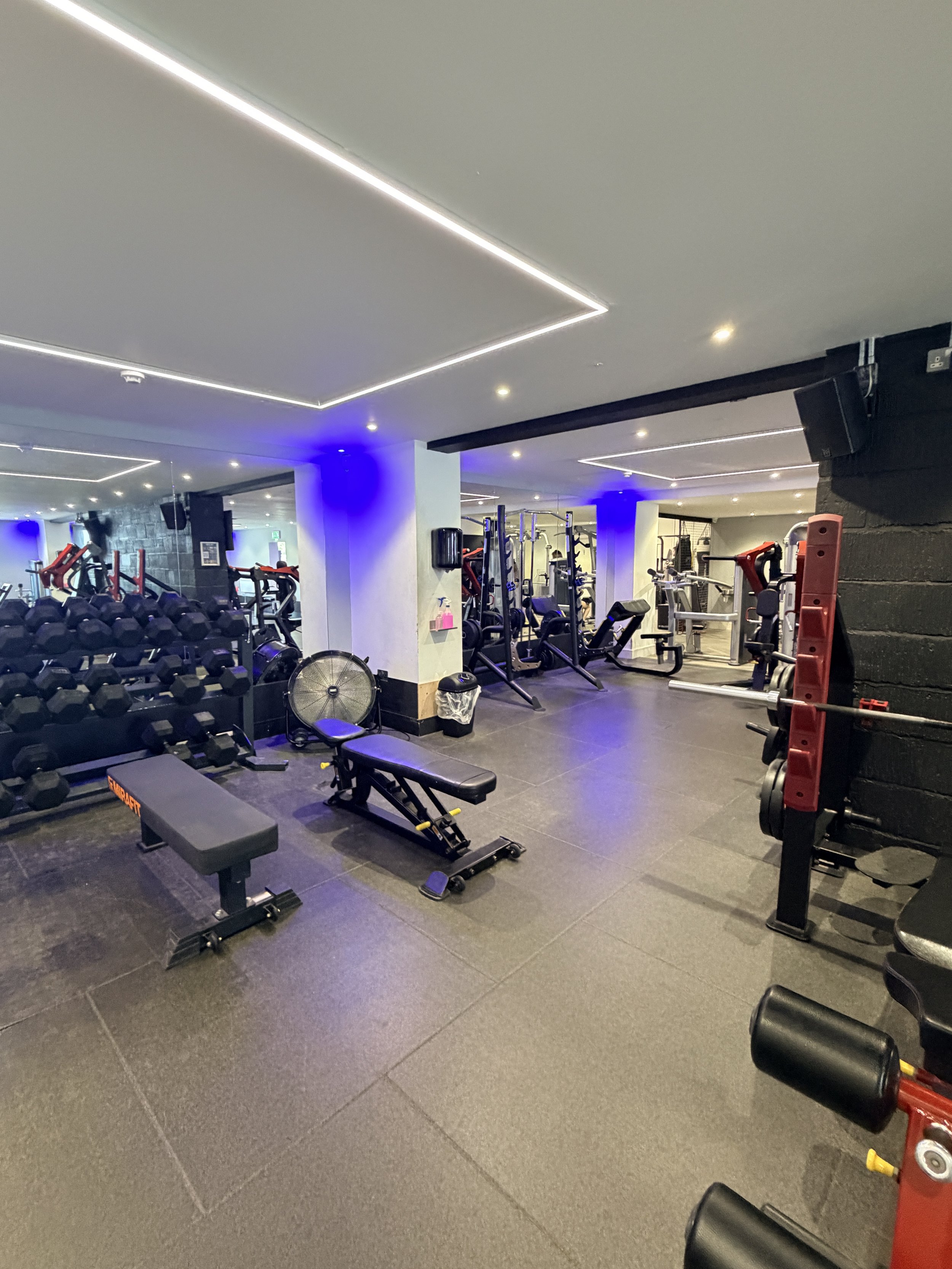 A gym with various workout equipment including dumbbells, benches, and resistance machines, under modern ceiling lighting with blue accent lights.