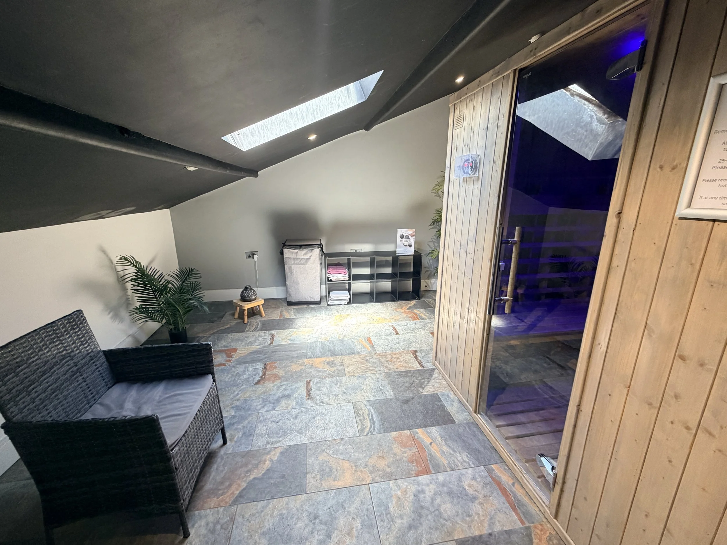 A room with slate flooring, a black wicker loveseat with gray cushion, a potted plant, a small table with a decorative jar, a shelving unit with towels, and a sauna with wooden exterior and glass door. Skylights with natural light and recessed ceilin