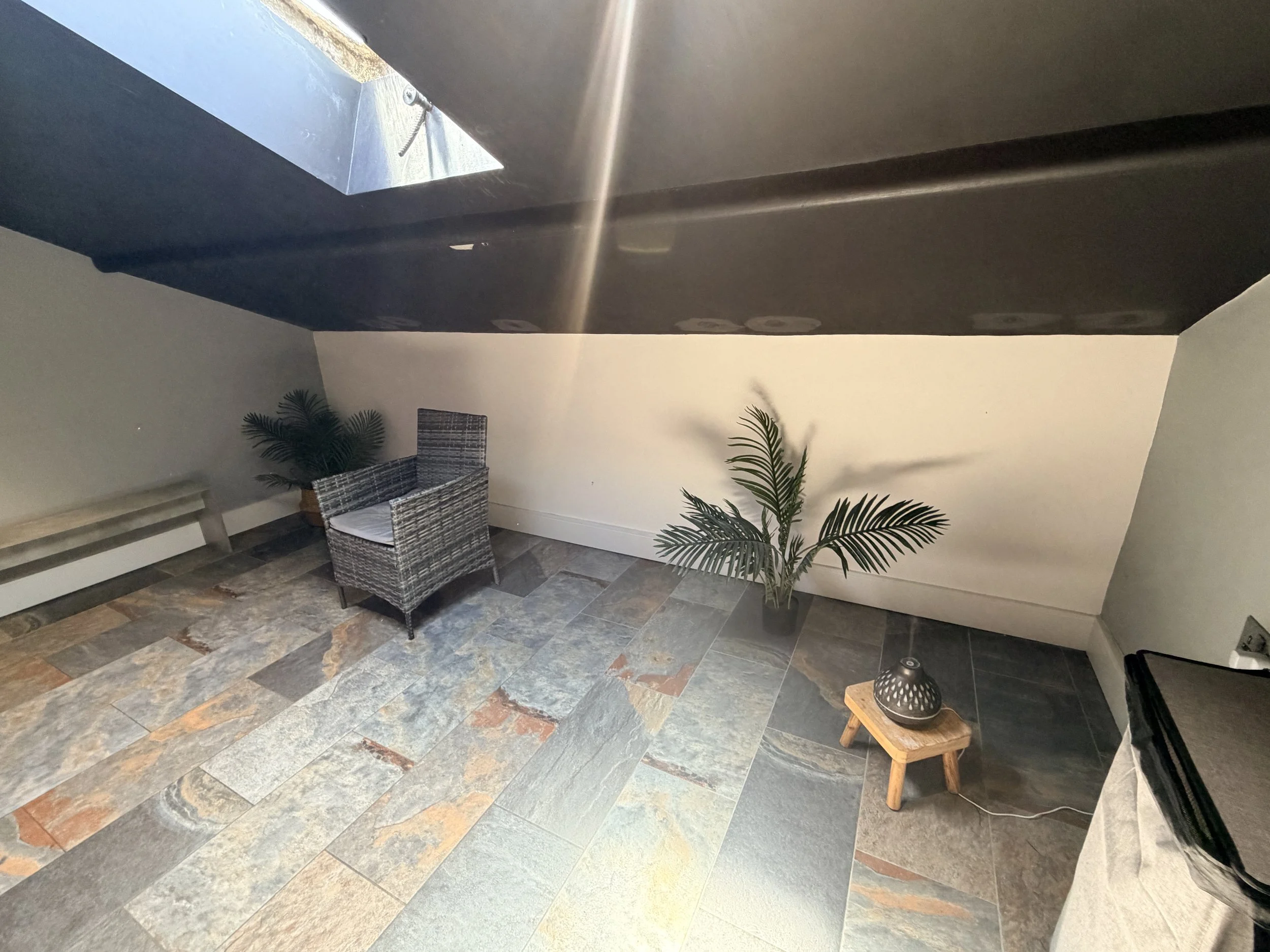 A cozy indoor space with a colorful brick-textured floor, a wicker chair with a plaid cushion, a small wooden side table with a decorative vase, and green plants in the background. There is also a section of a wall heater and a black office chair.