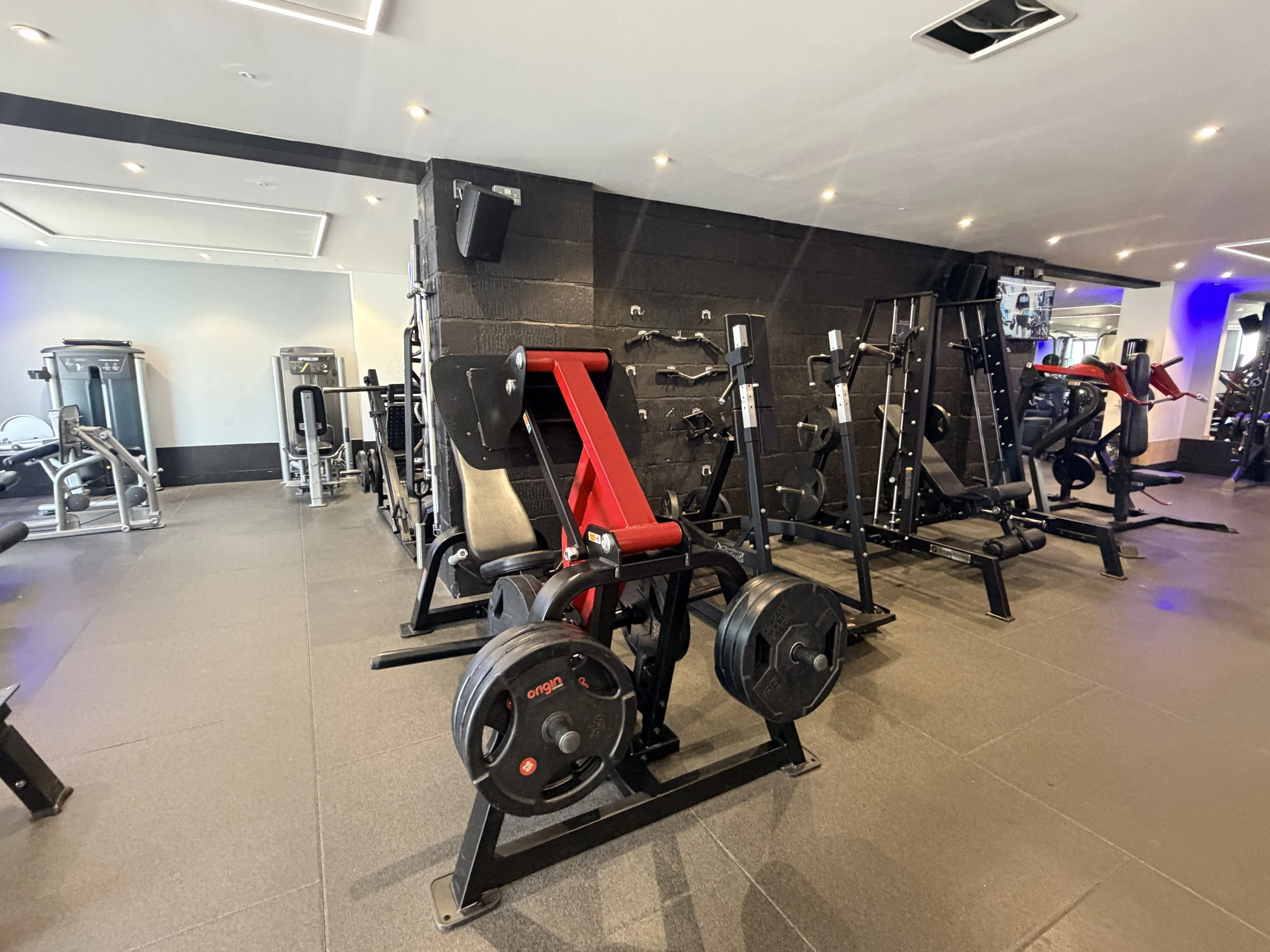 Gym equipment including barbells, weight plates, and various workout machines in a modern gym with gray flooring and a black accent wall.