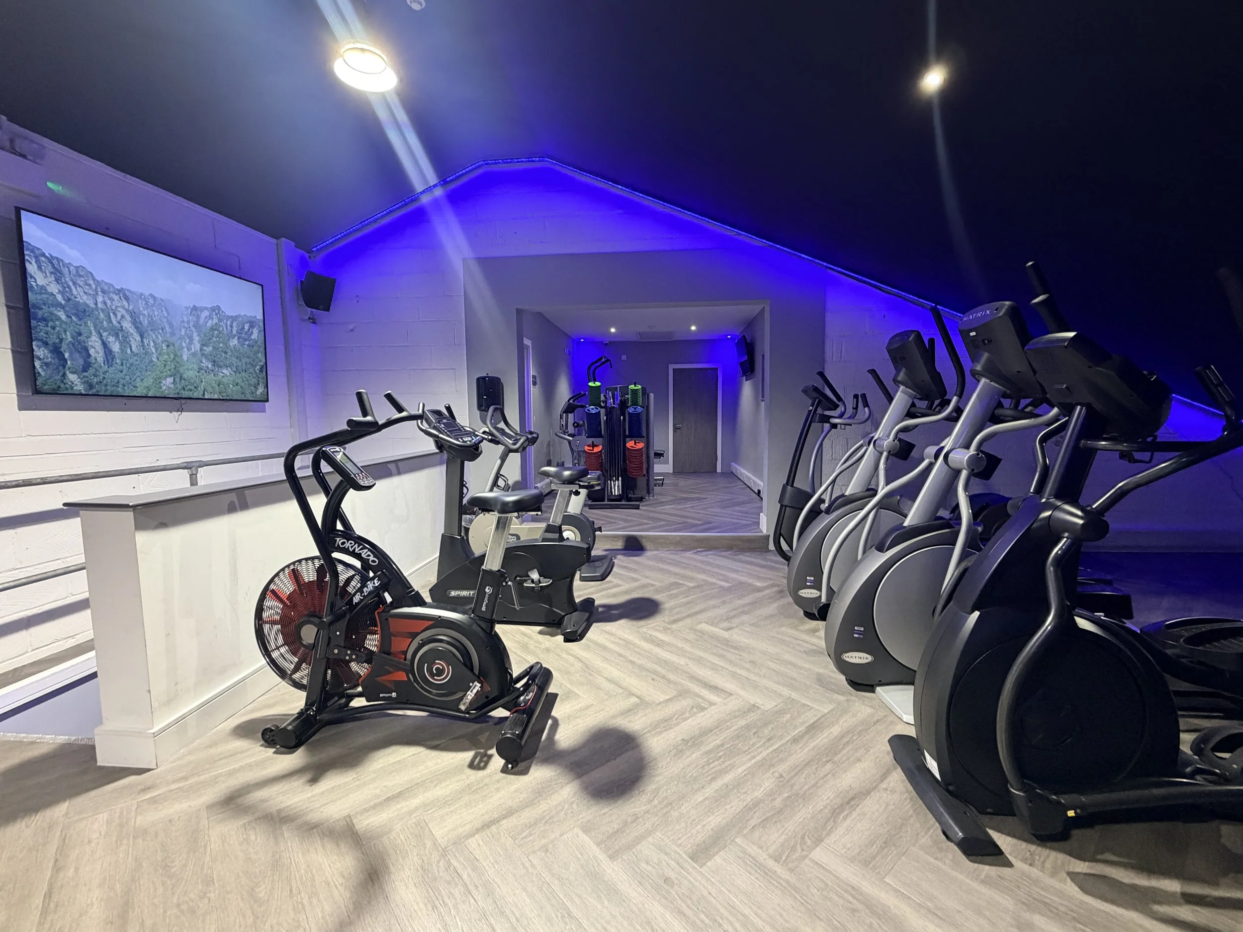 Indoor gym with exercise machines including elliptical trainers and a stationary bicycle, blue accent lighting, a large wall-mounted screen, and beige wood flooring.