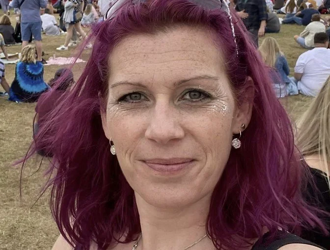 Close-up of a woman with purple hair outdoors at a crowded event, smiling at the camera.