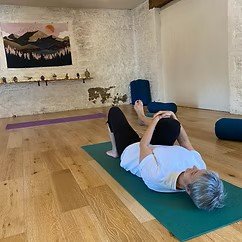 An person doing a stretching exercise on a yoga mat in a room with wooden floors, a painting on the wall, and exercise equipment in the background.