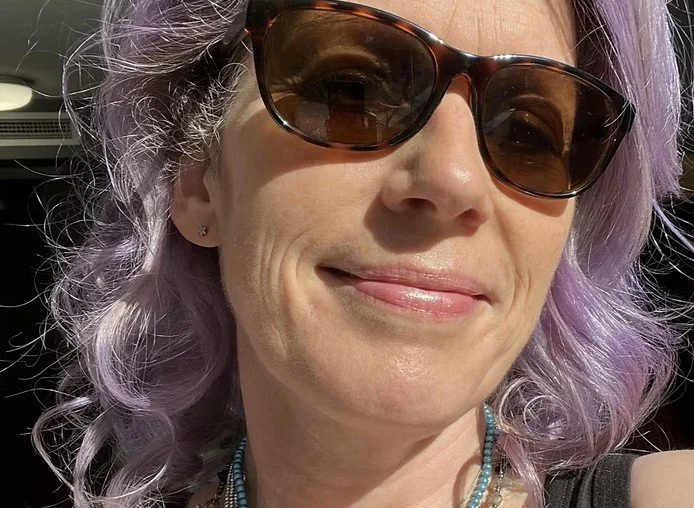 A woman wearing sunglasses with purple hair, smiling.