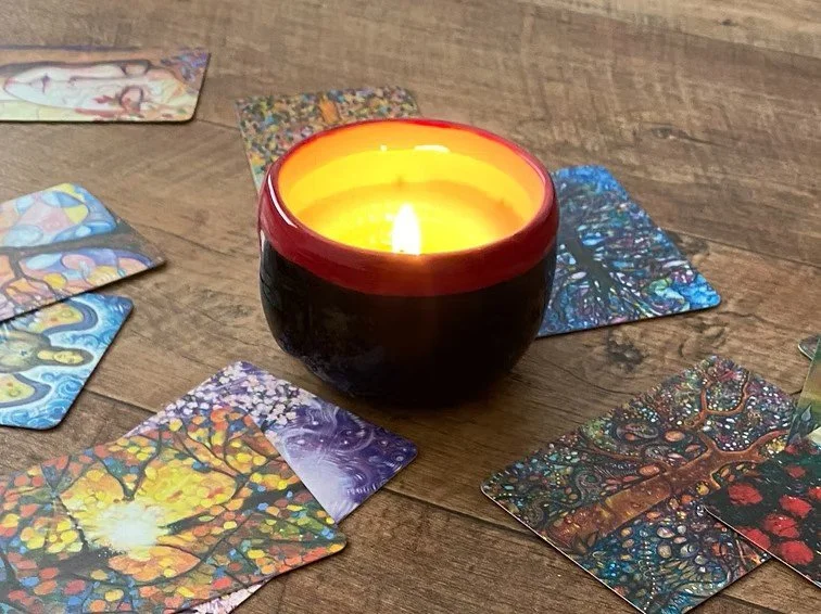 A lit candle in a black and red ceramic holder on a wooden surface, surrounded by colorful tarot or oracle cards with intricate, vibrant artwork.