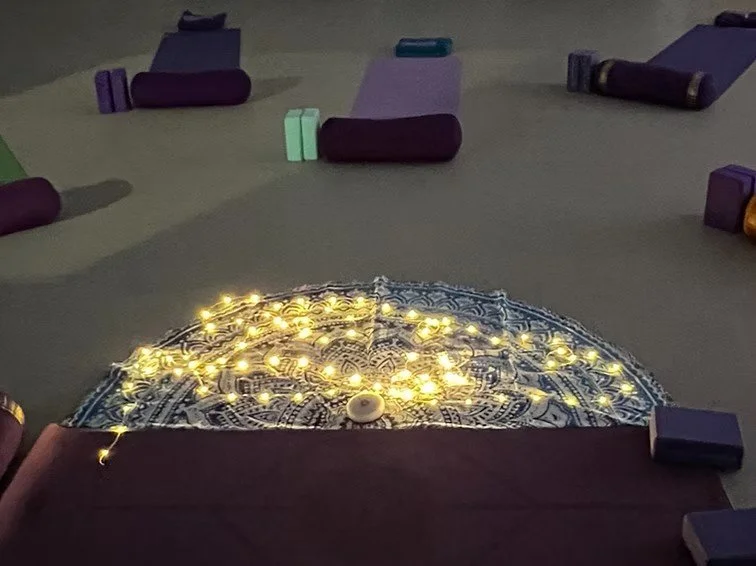 A yoga mat with string lights on top, surrounded by folded yoga mats in various colors in a dimly lit room.