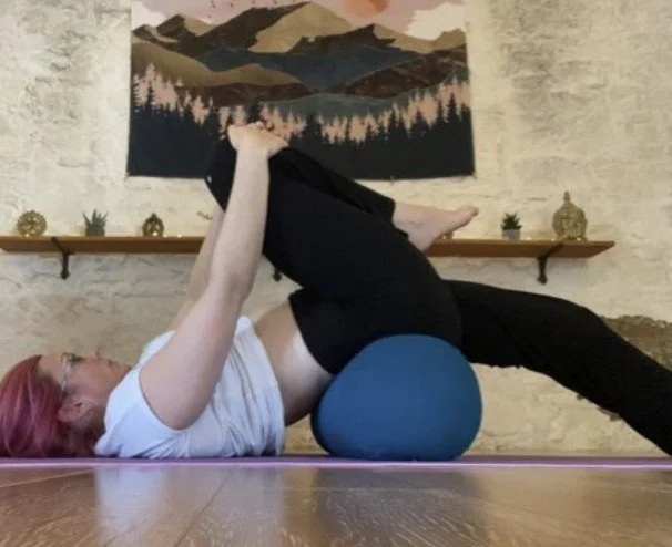 A woman lying on her back on a yoga mat, using a foam roller under her hips, practicing a stretching or exercise pose in a room with a decorative wall and a mountain landscape painting