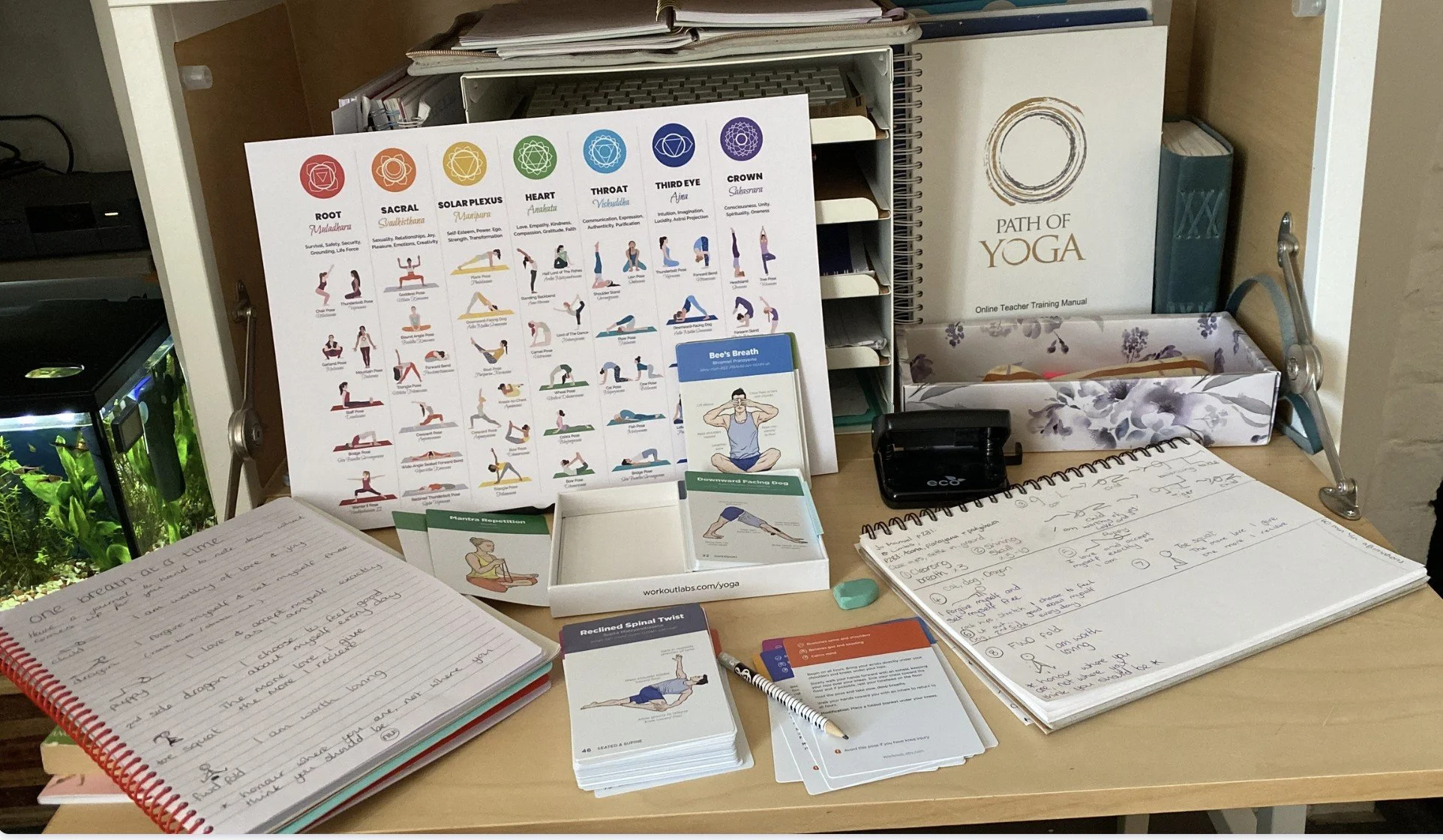 Desk with notes, books, yoga cards, and a yoga poster. Notebooks and writing utensils are scattered across the surface.