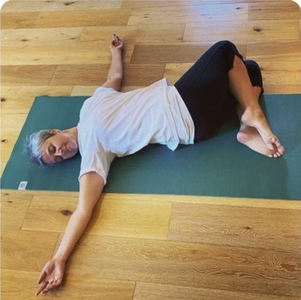 A woman lying on her side on a yoga mat in a relaxed pose on a wooden floor.