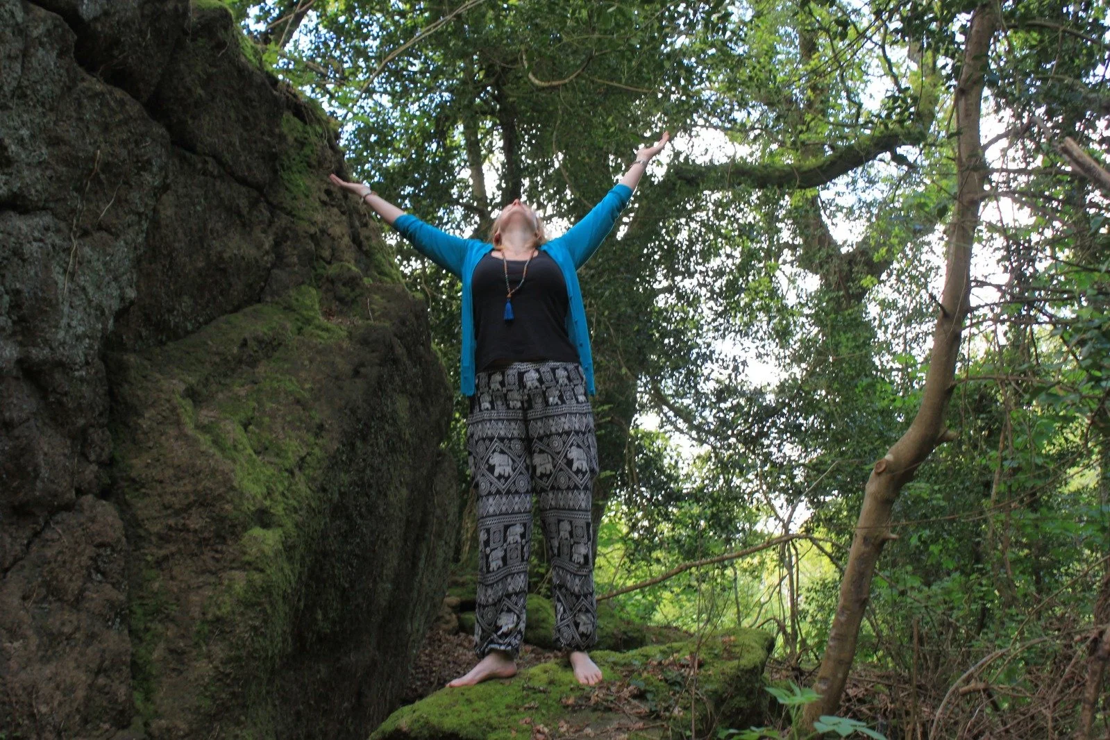 A woman standing outdoors in a green forest, with arms outstretched and head tilted back, expressing joy or freedom.