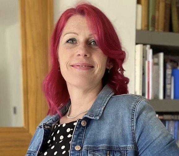 A woman with shoulder-length red hair, wearing a denim jacket over a black and white polka dot top, standing in front of a bookshelf.