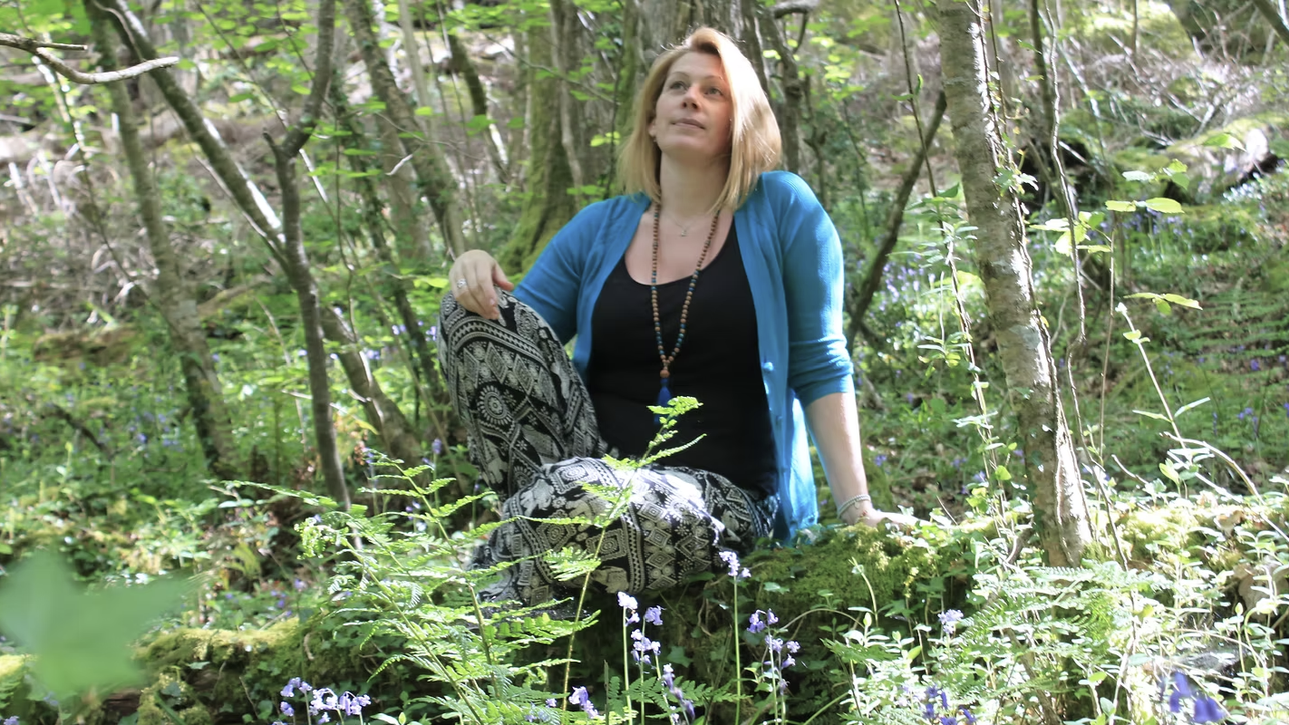 A woman sitting among trees and greenery in a forest, wearing a blue cardigan, black top, and patterned pants, looking upward with a peaceful expression.