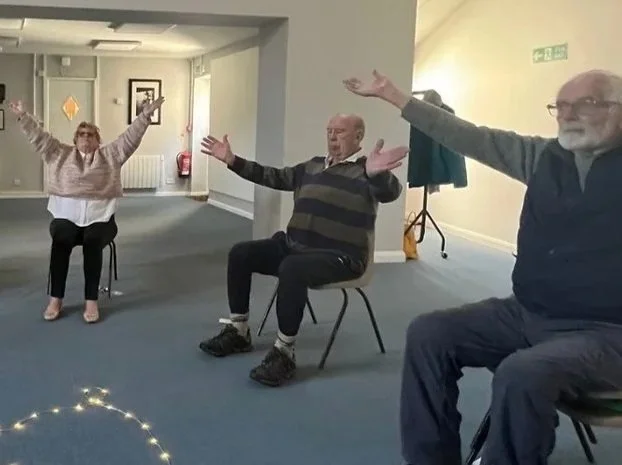 Three people are sitting on chairs in a room, with their arms raised, participating in an activity. The woman on the left is wearing glasses, a light-colored sweater, and black pants. The man in the middle is wearing a striped sweater and dark pants. The man on the right is wearing glasses, a dark shirt, and jeans. There are fairy lights on the floor in the foreground.