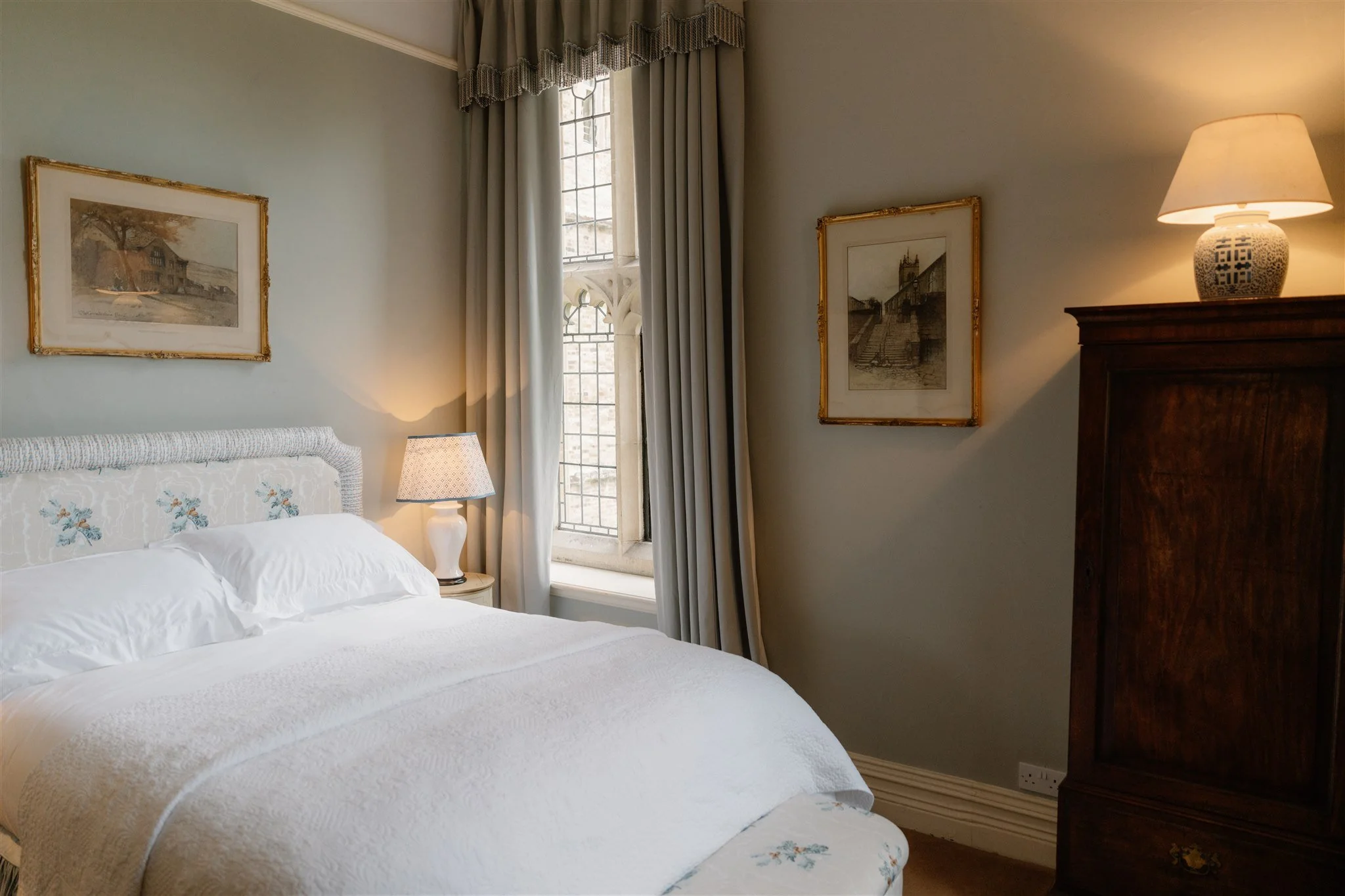A cozy bedroom with a bed, white quilt, and floral pillow. There's a bedside lamp, framed artwork on the wall, and a window with curtains letting in natural light.