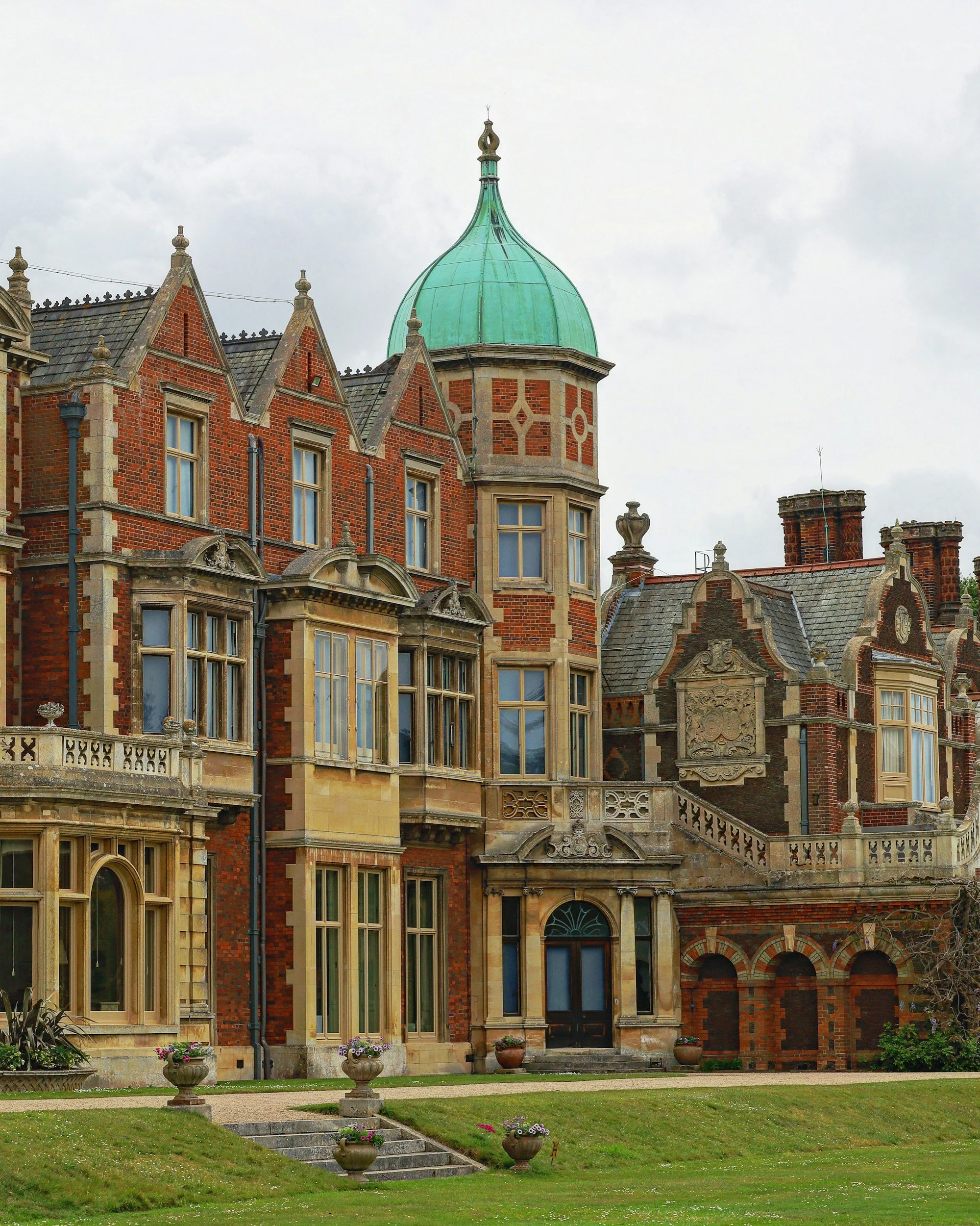 Front view of a large historic brick mansion with multiple pointed gables, decorative stone detailing, and a green domed turret under a cloudy sky, with a well-maintained lawn and flower pots in the foreground.