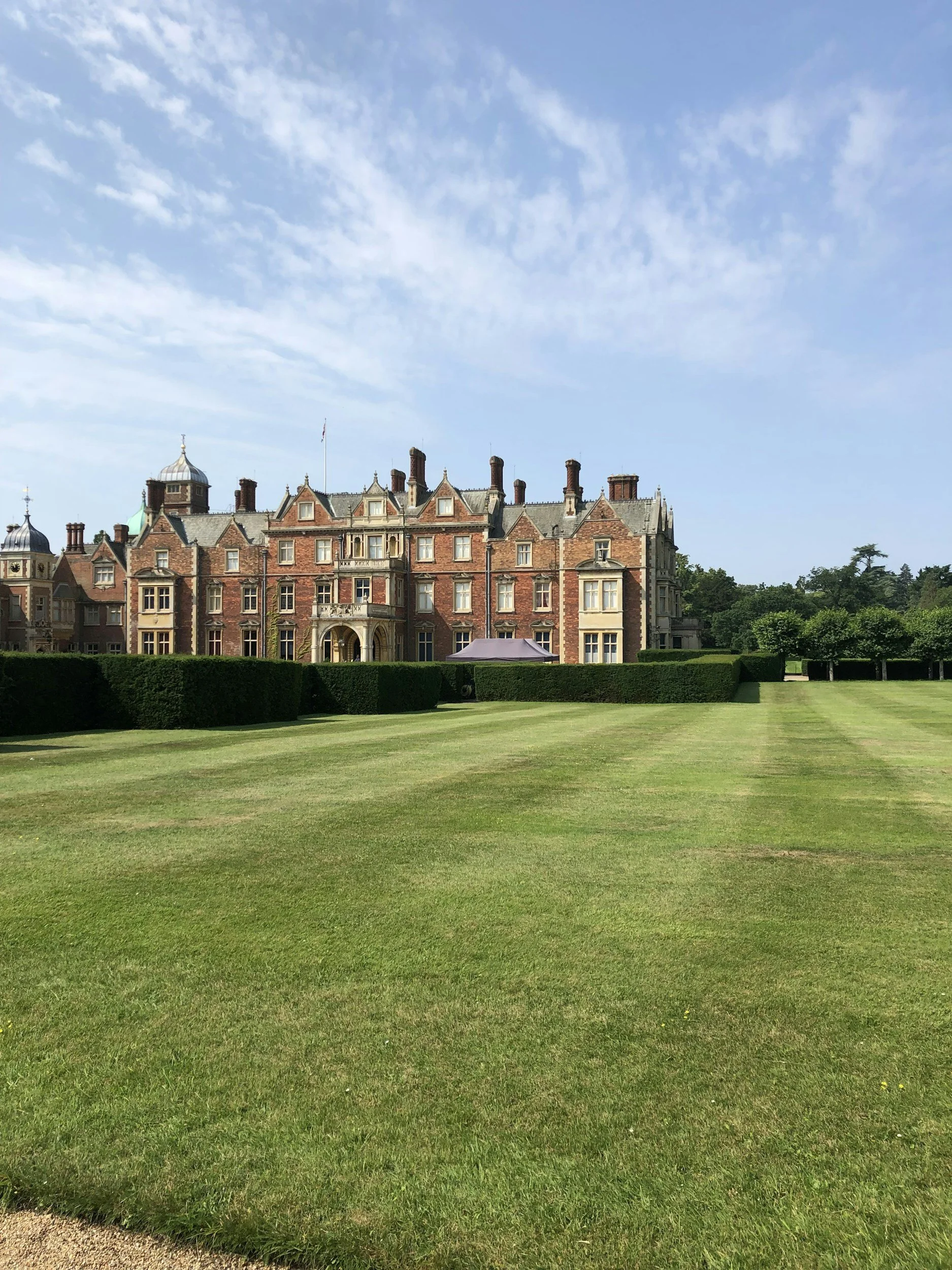 A large, historic brick mansion or estate with a tower and decorative roof features, set against a backdrop of green trees. The foreground shows a well-manicured lawn and a gravel pathway.