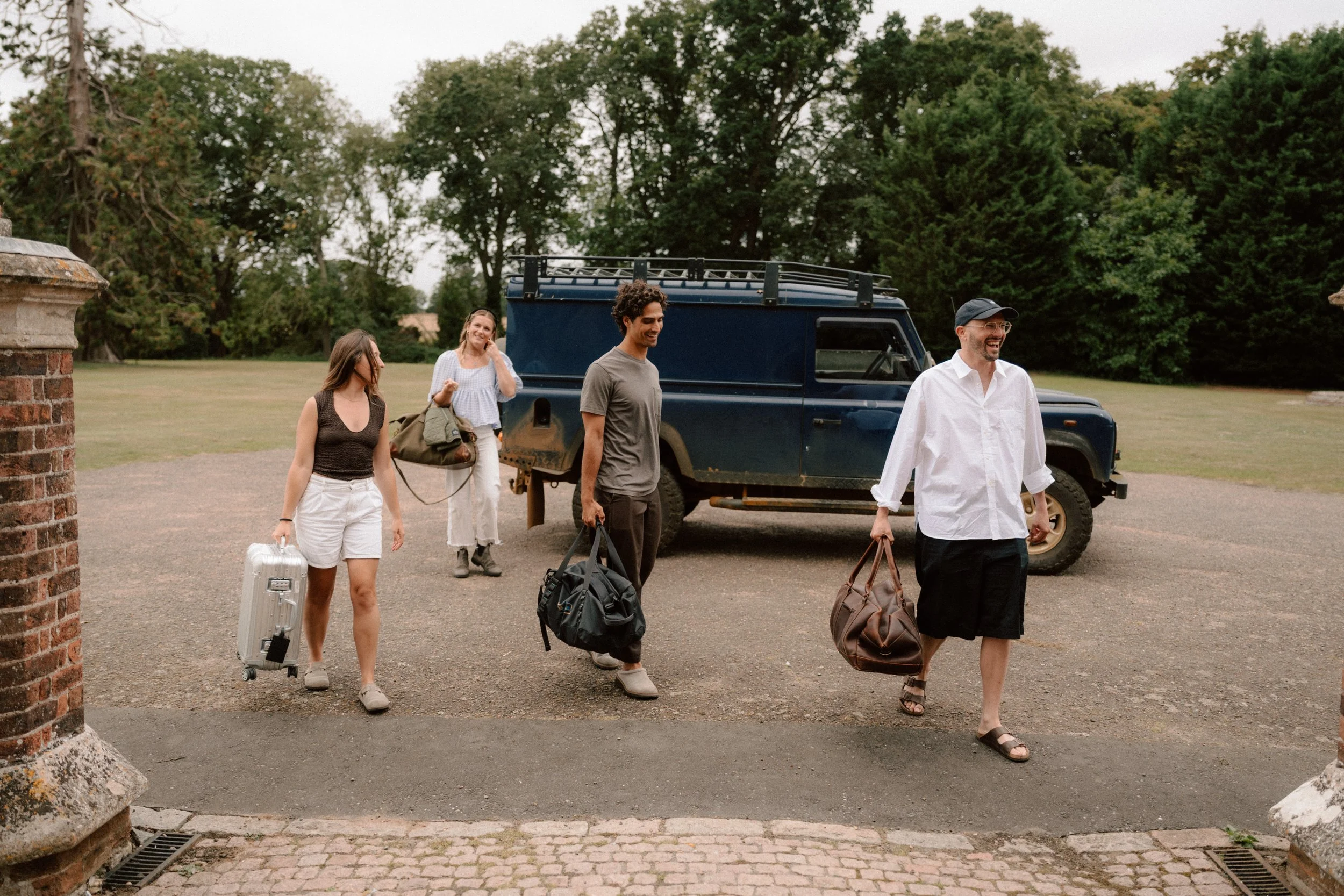 Four people walking with luggage away from a dark blue off-road vehicle in a park-like area with large trees and a cloudy sky