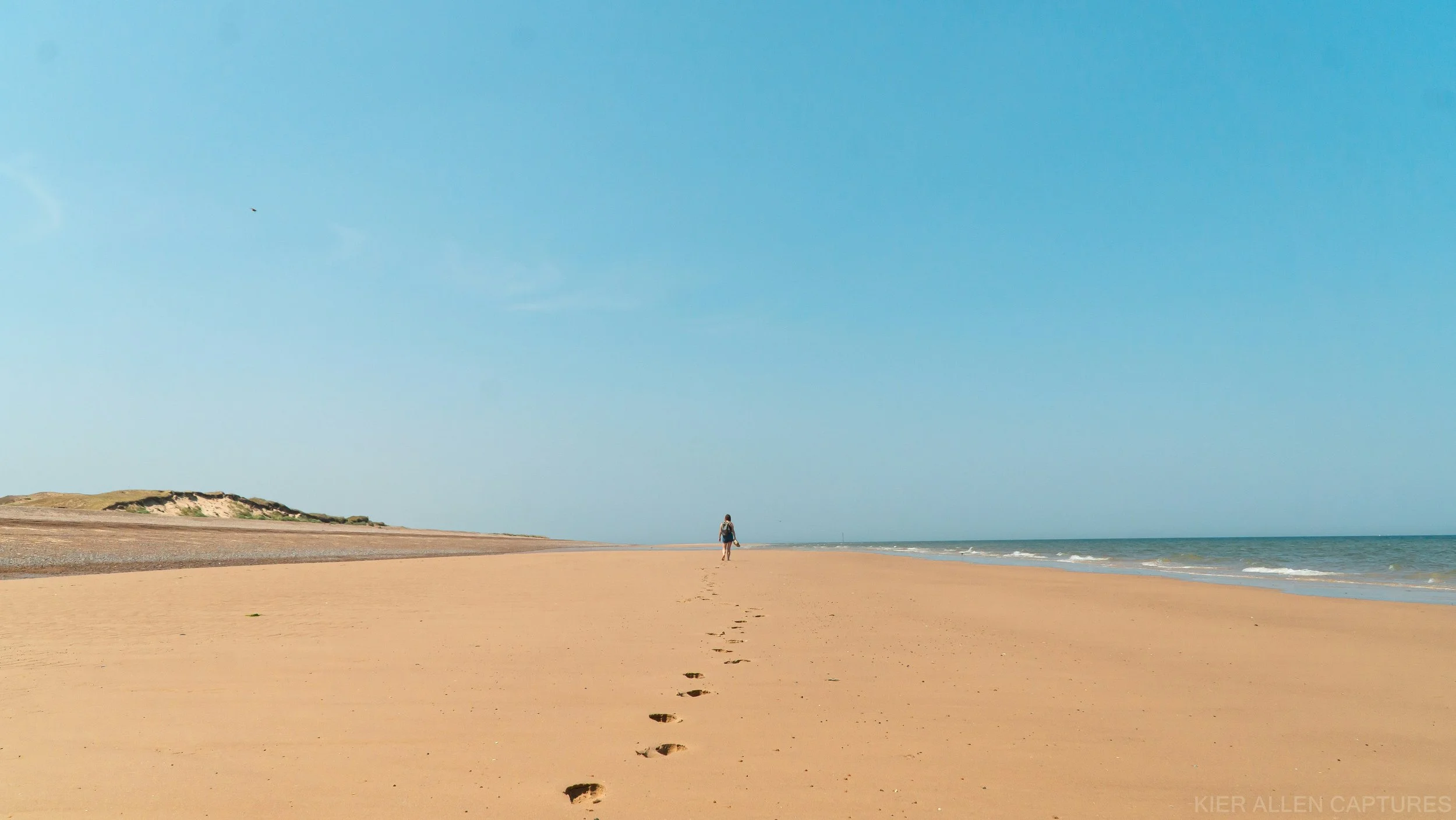A person walking along the beach, leaving footprints in the sand, with the ocean and a clear blue sky in the background.