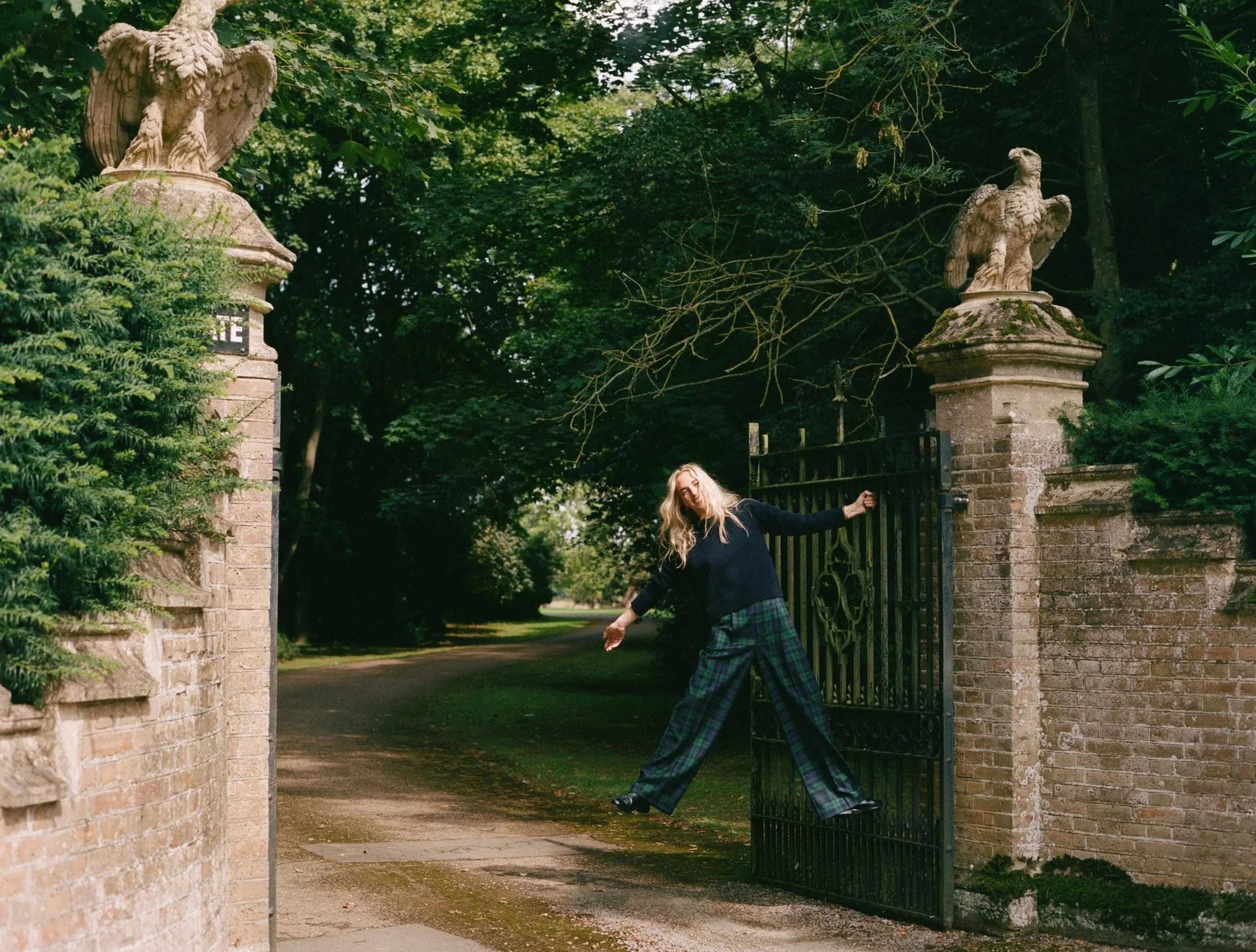 Woman with long blonde hair wearing a dark top and traditional tartan pants opening a black gate in a green park with stone statues and lush trees.