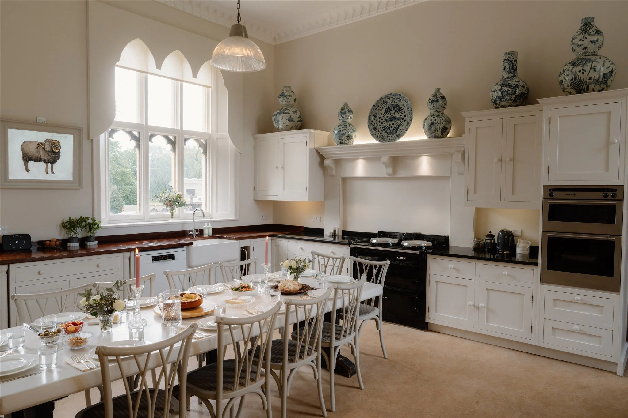 A bright kitchen with a large window, white cabinets, a black stove, and a dining table set for a meal with candles and flowers, decorated with blue and white vases on top of the cabinets.