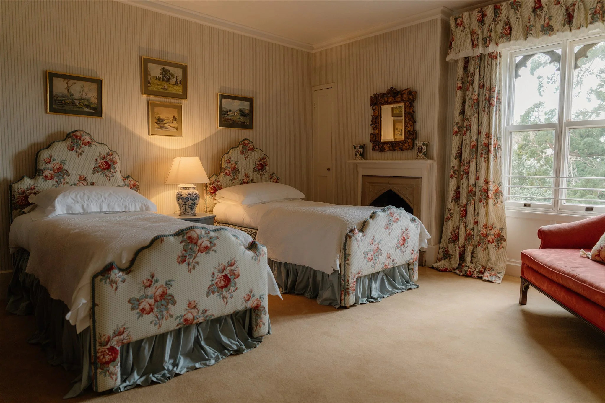 A cozy bedroom with two beds that have floral patterned headboards, a bedside lamp, a mirror, and a window with floral curtains showing trees outside.