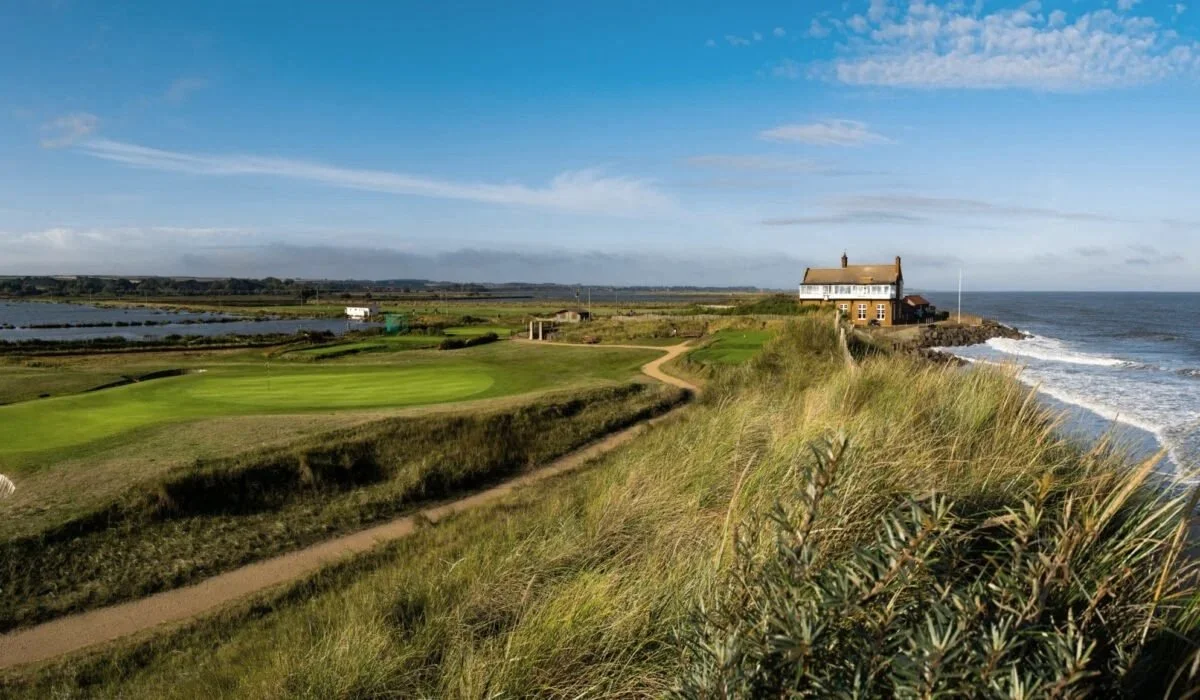 Scenic coastal landscape with a golf course, grassy dunes, a building on a cliff near the ocean, and a clear blue sky.